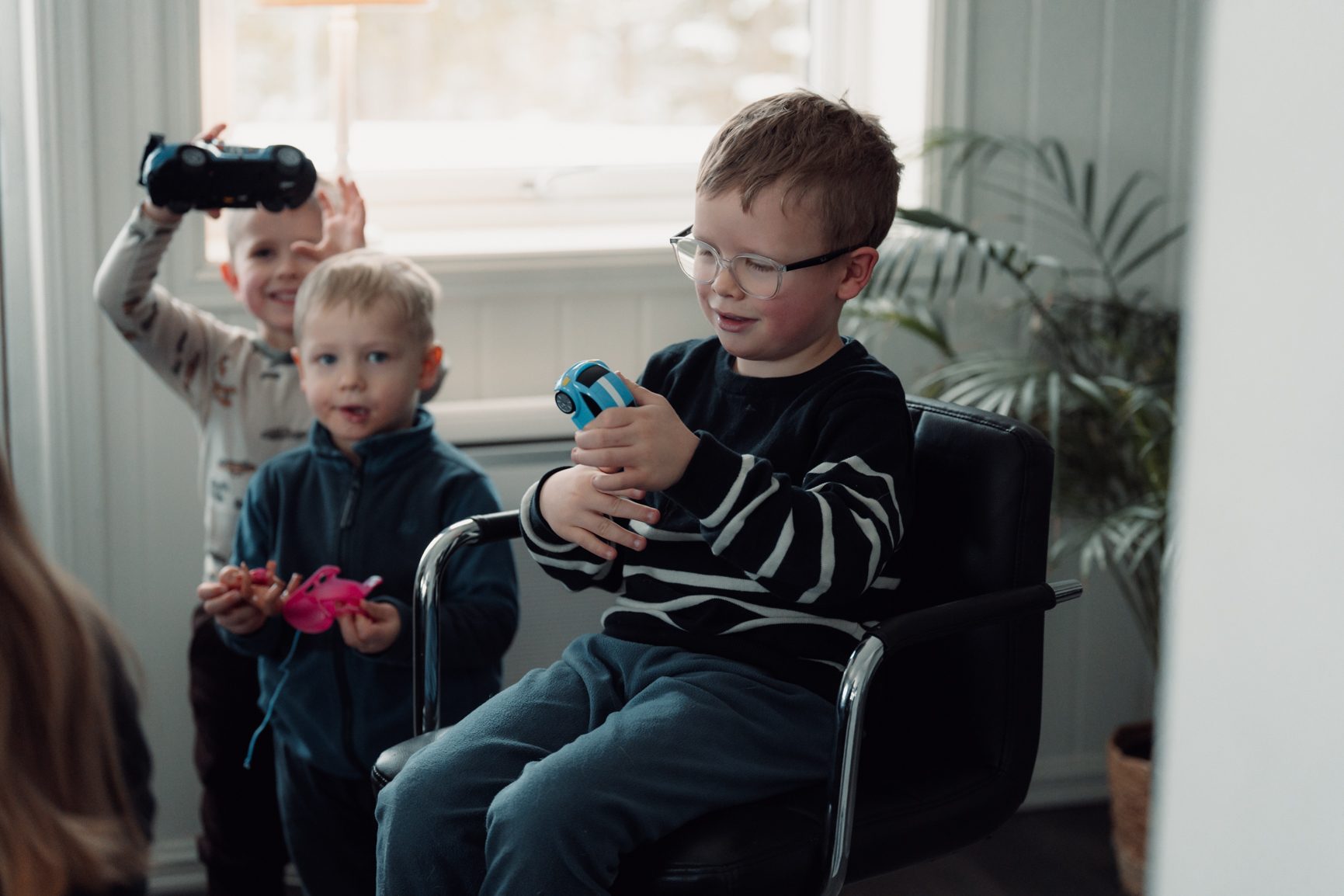 Three young boys playing with toy cars: one seated with a blue car, one standing with a pink toy, and one in back.