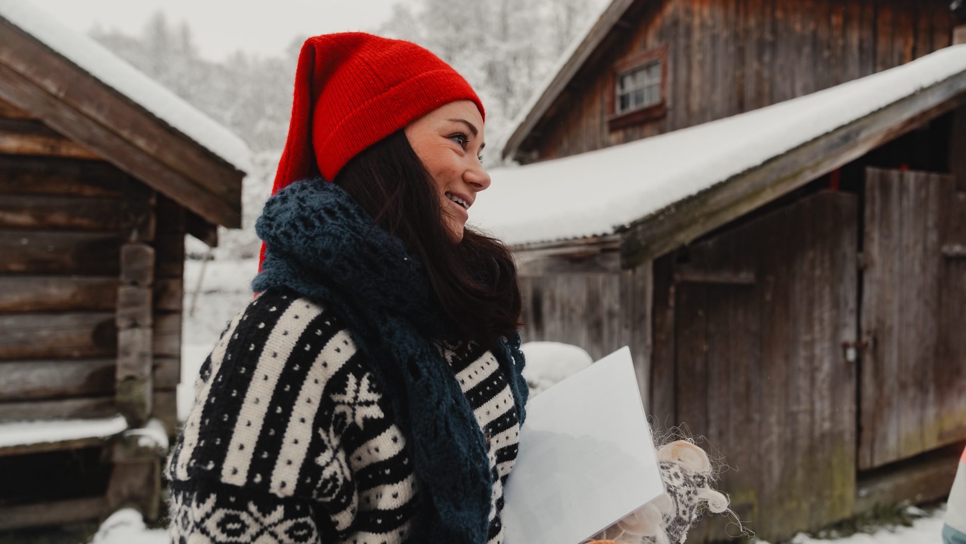 Smiling woman in red hat and winter clothes in snowy village with wooden cabins.