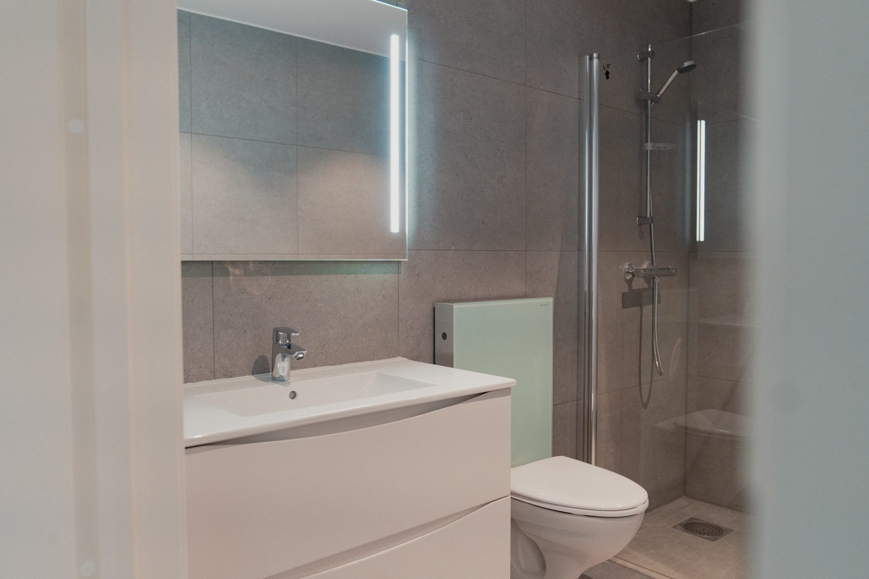 A modern bathroom with a light grey tiled wall, white sink vanity, toilet, illuminated mirror, and glass shower enclosure.