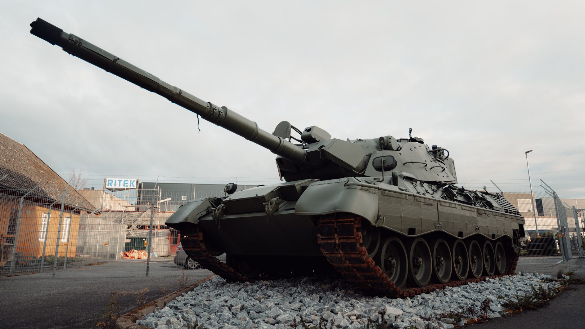 A large green military tank with a raised gun barrel sits on rocks, industrial buildings in background.