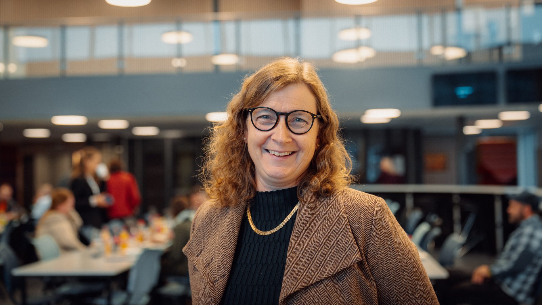 Smiling woman with glasses, curly brown hair, brown coat, and gold chain in an indoor setting.