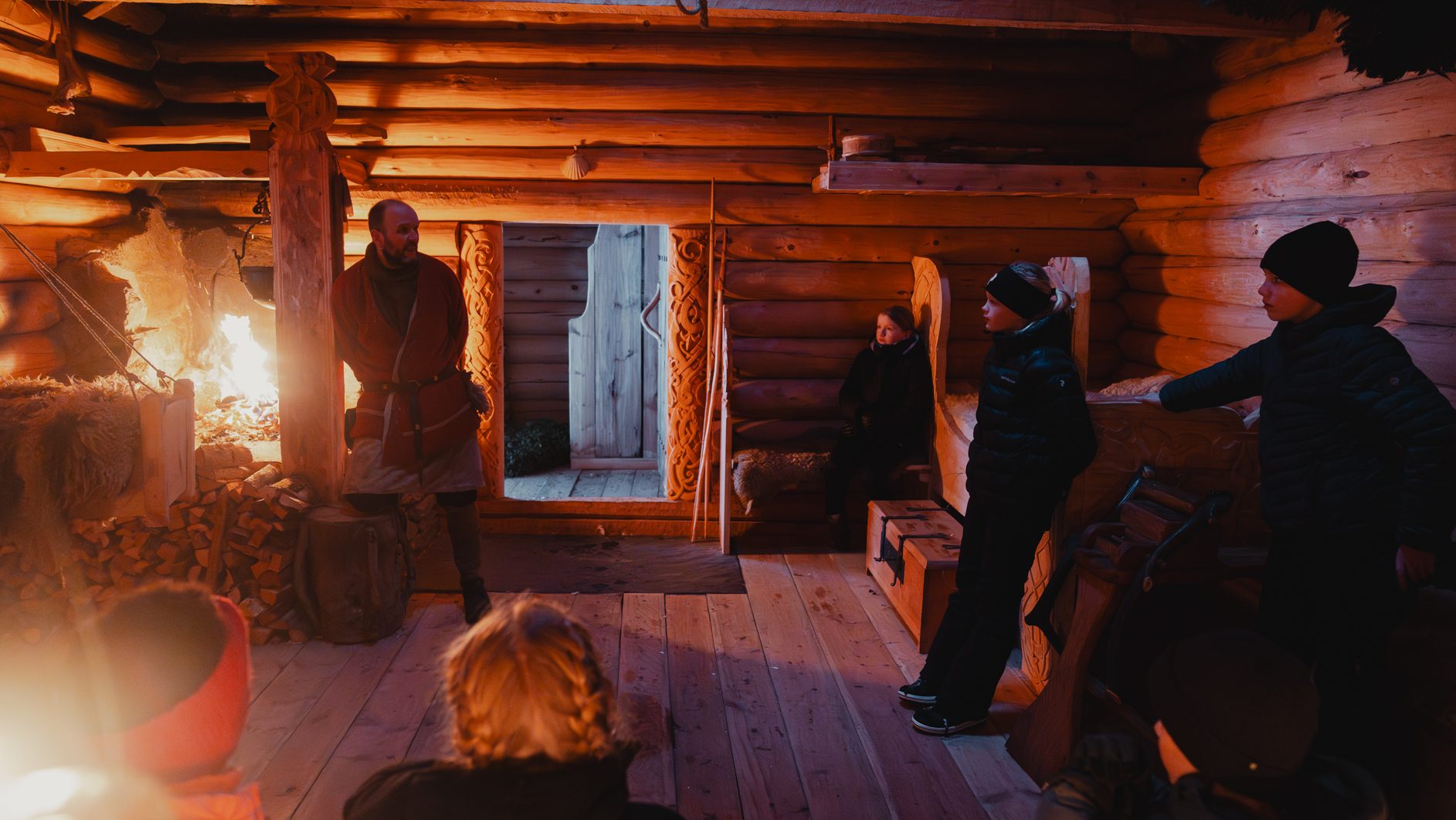 Man in period costume by a roaring fire in a wooden cabin, people watching, low light.