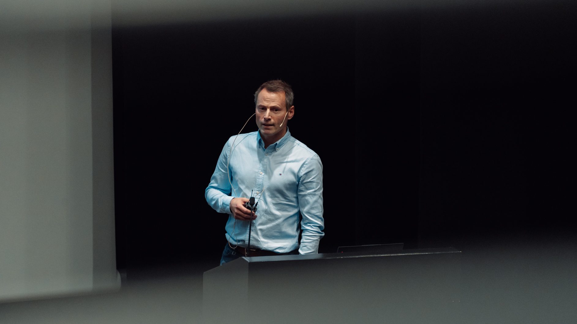 A man in a blue shirt speaks into a headset mic on a dark stage, standing at a podium.