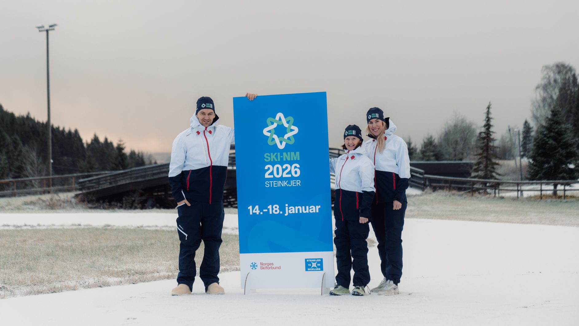 Three people in winter gear holding a blue "SKI-NM 2026 Steinkjer" sign in the snow.