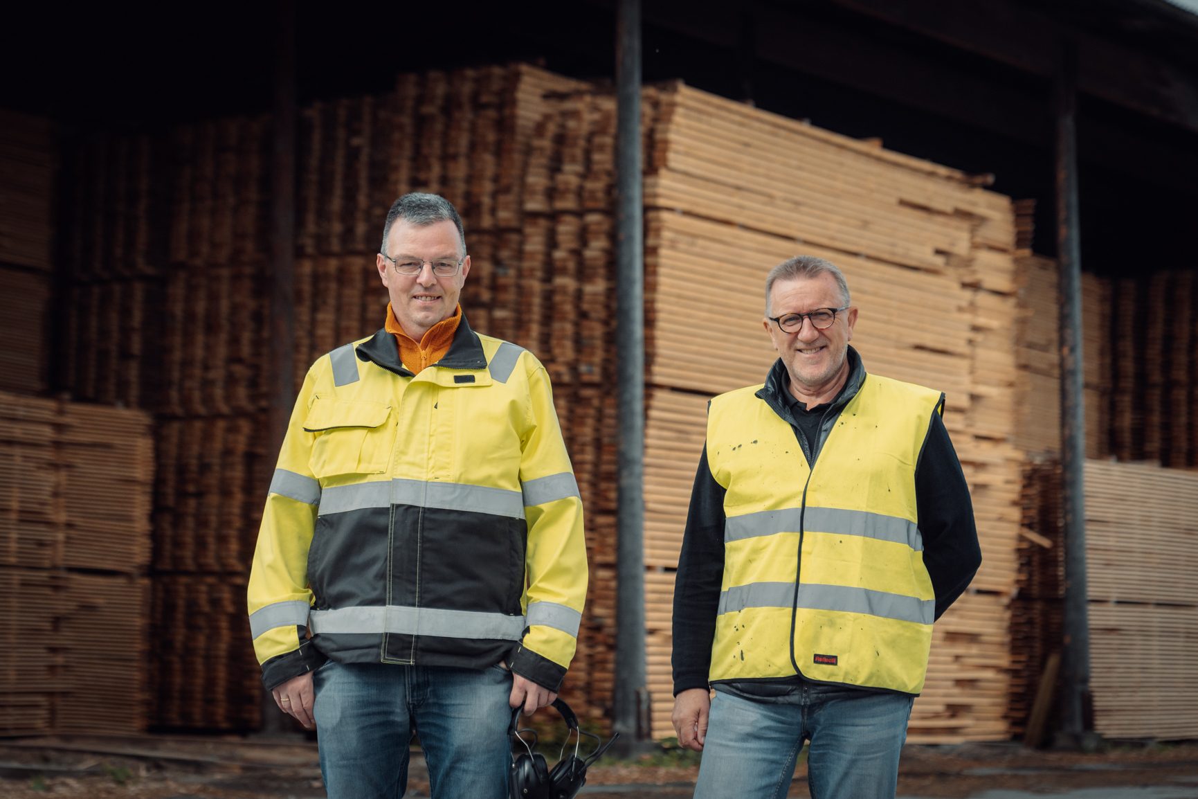 Two men in yellow safety vests standing in front of stacked lumber.