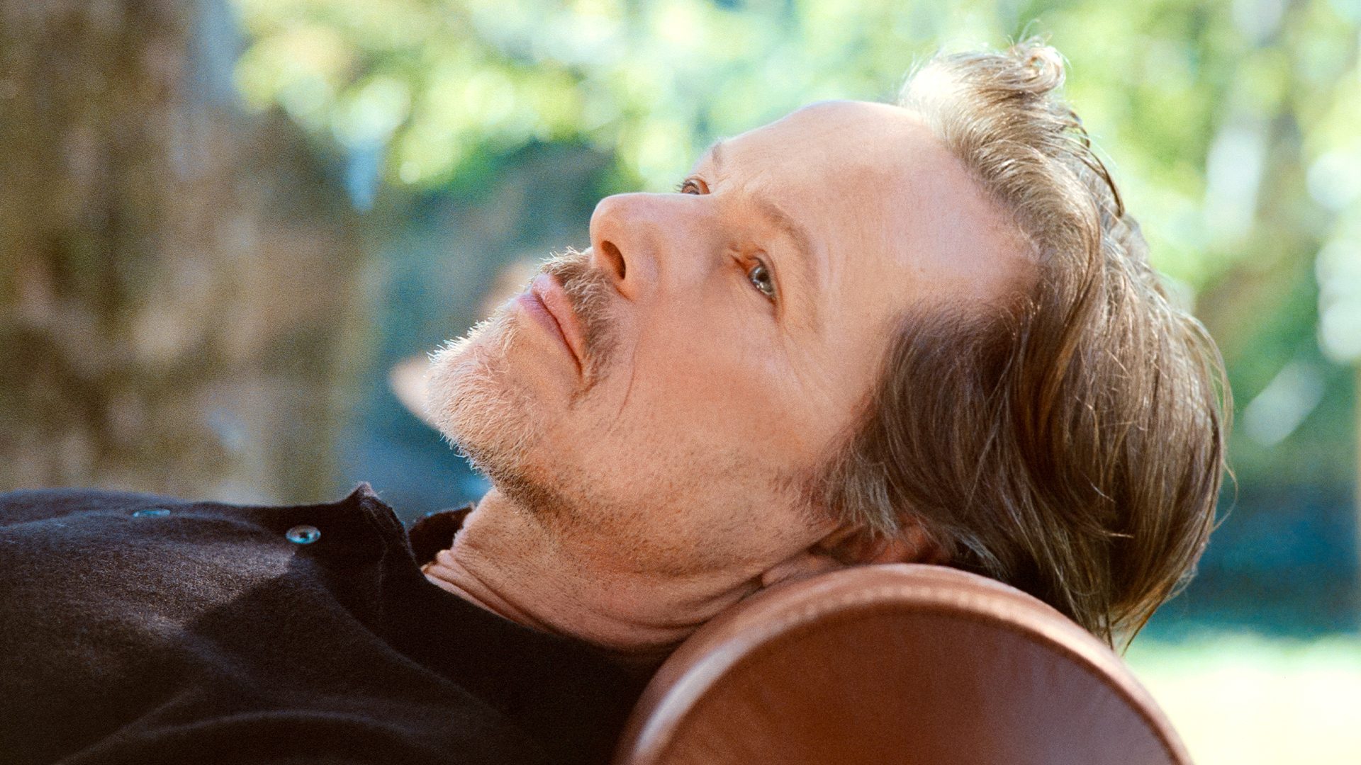 A man with a beard and brown hair lies back, looking up pensively, blurred green background.