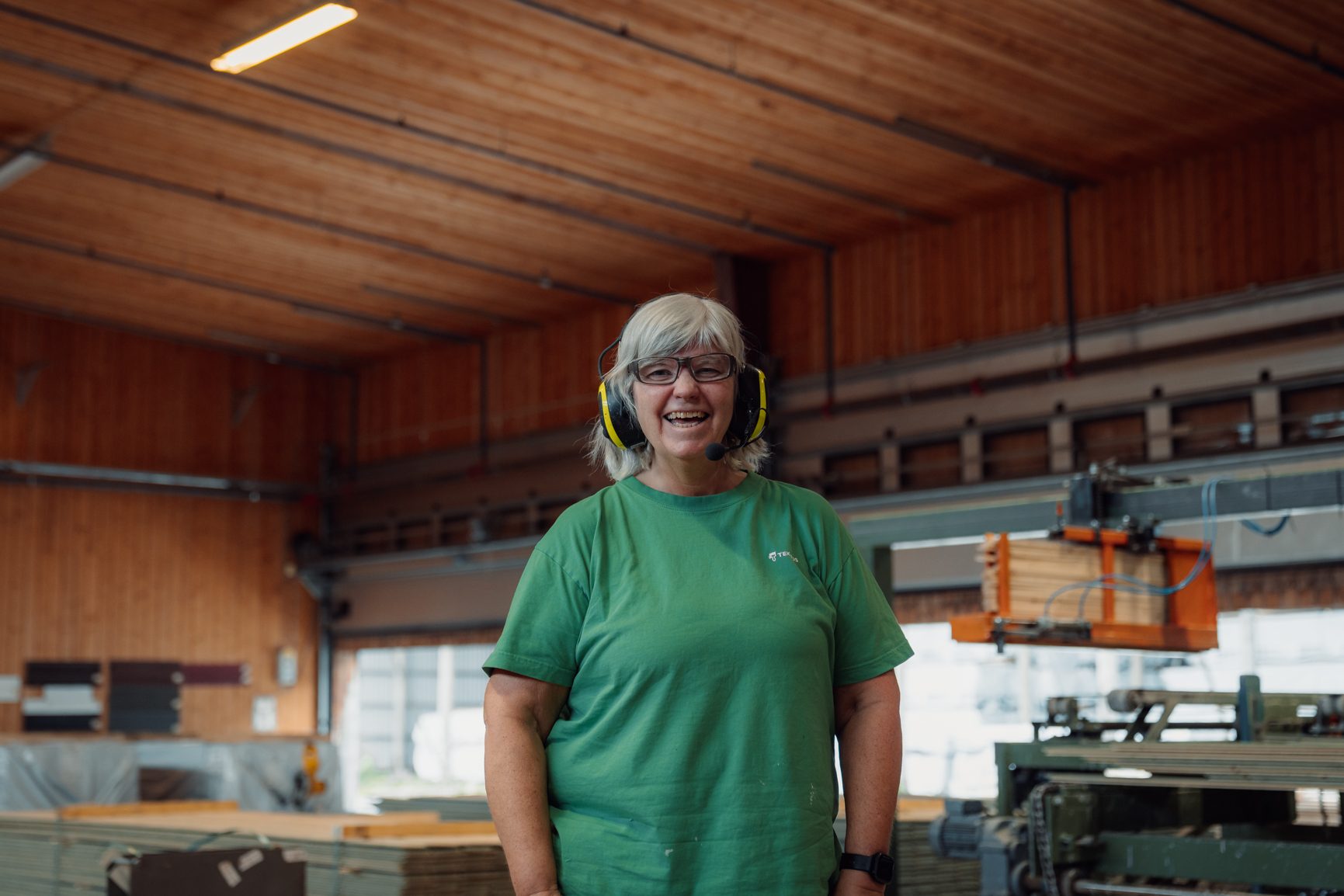 Smiling older woman with gray hair, headphones, green shirt in a workshop.