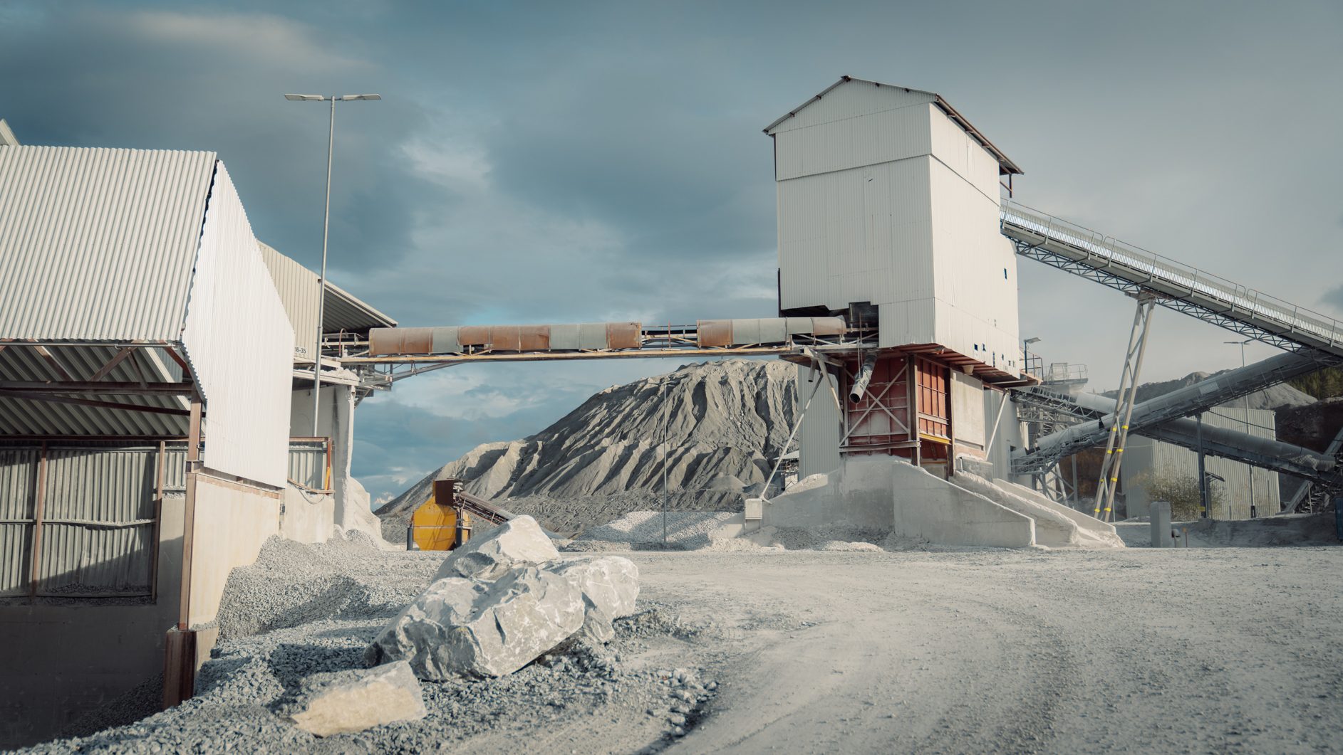 Industrial quarry with white buildings, conveyor belts, and large piles of crushed rock under a cloudy sky.
