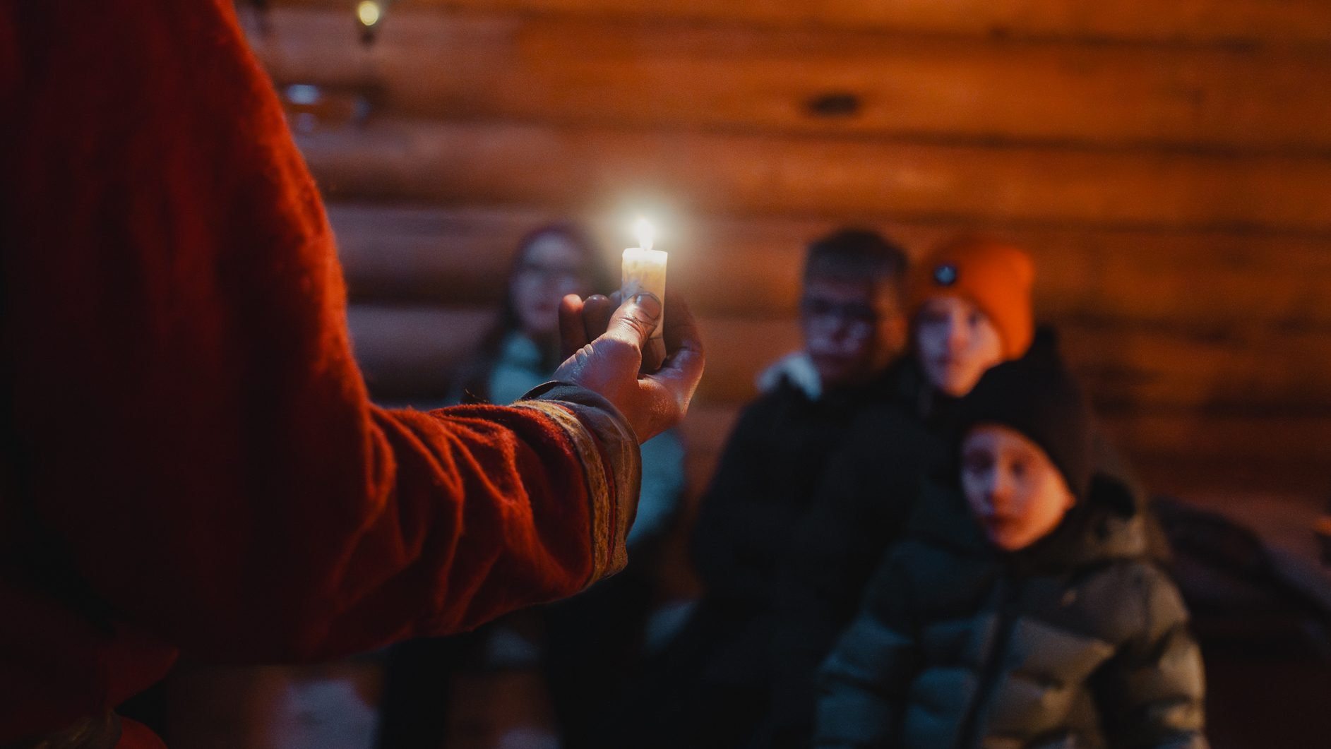 A hand holds a lit candle, illuminating blurred faces in a dark wooden interior.