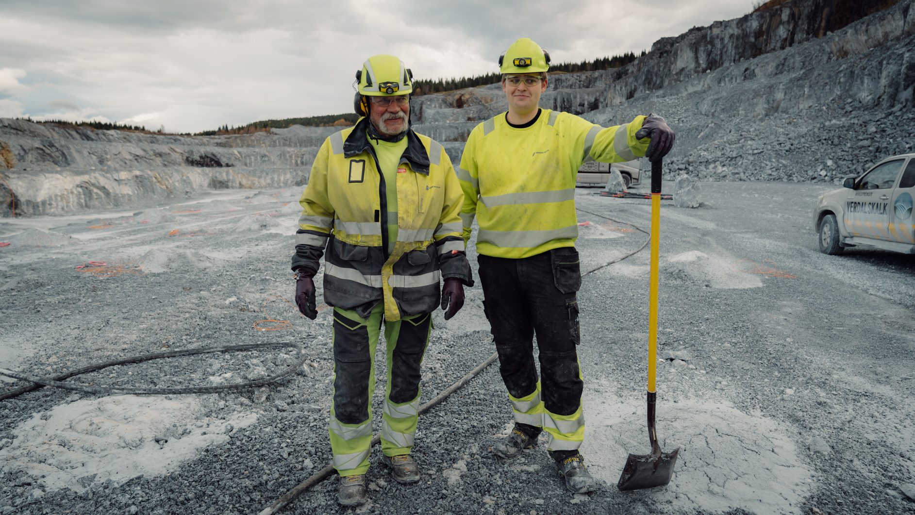 Two men in safety gear stand in a quarry, one with a shovel.