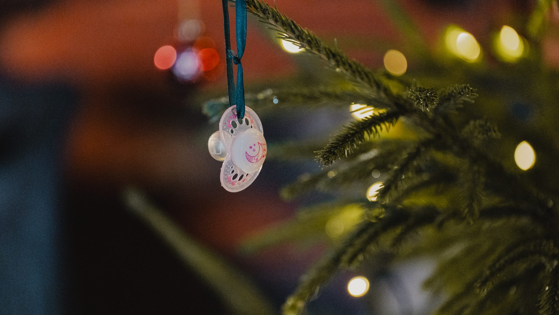 A baby pacifier hangs as an ornament on a Christmas tree, with blurred lights in the background.