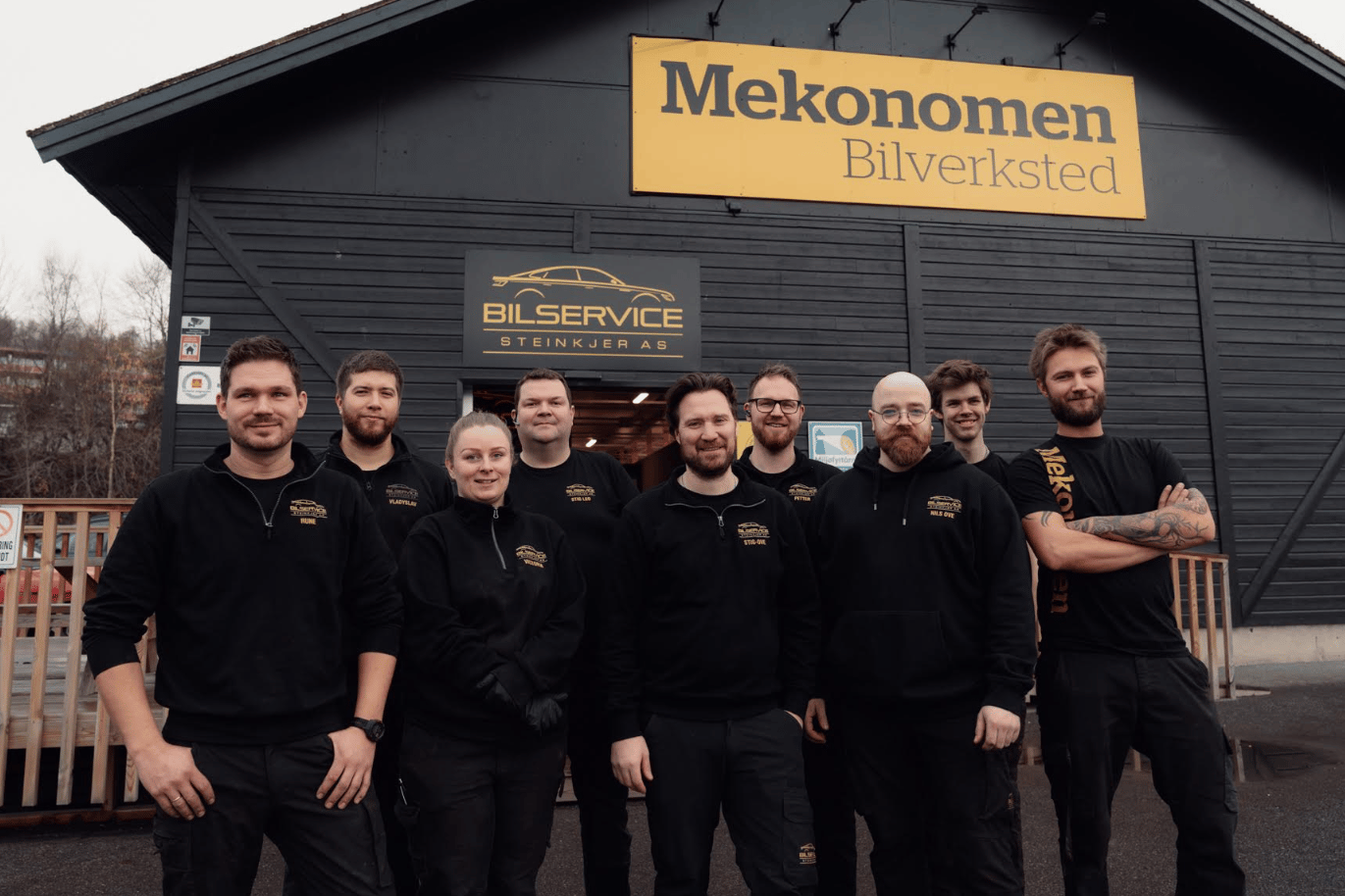 A smiling team of mechanics in black uniforms stands in front of their auto repair shop.