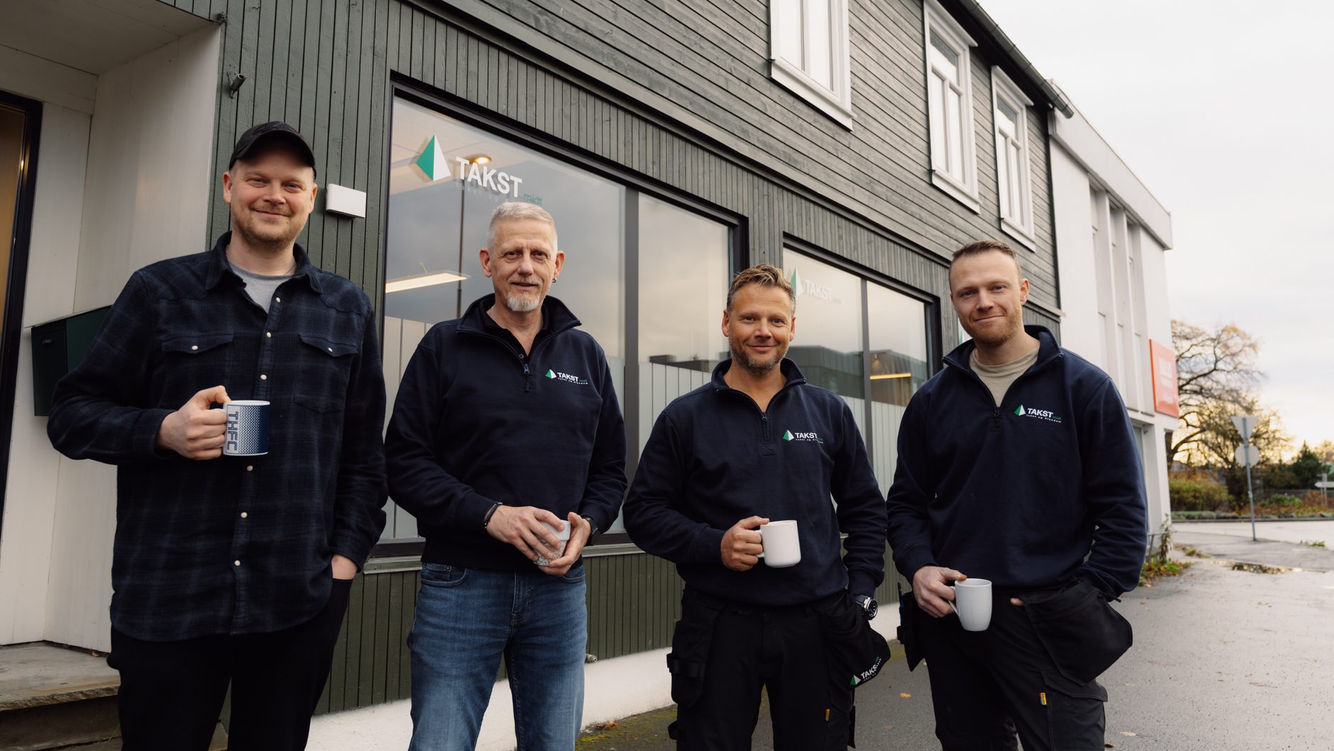 Four men holding mugs outside a building with the "TAKST" logo.