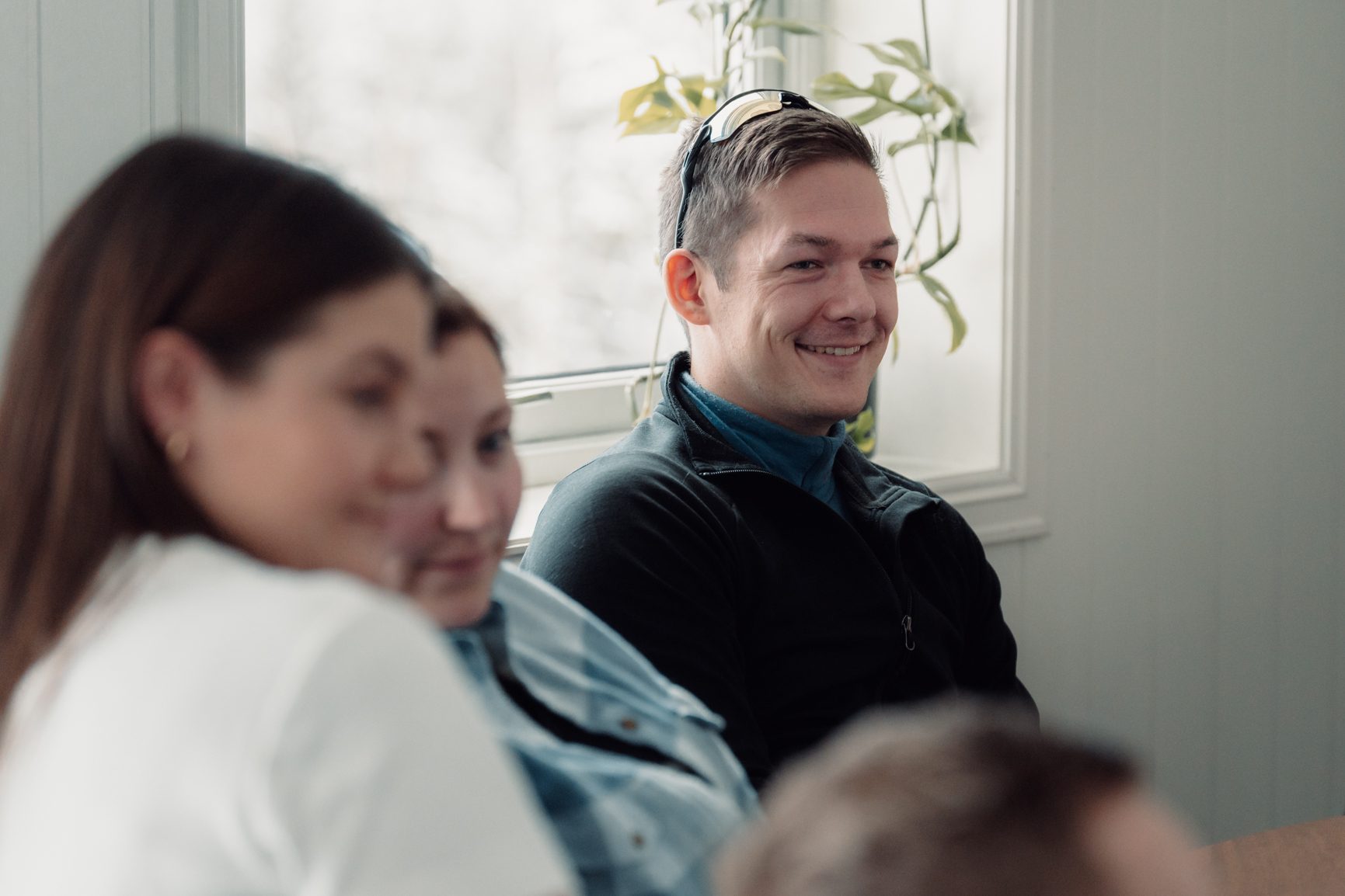 A smiling man with glasses on his head looks right while sitting with other people, blurred in the foreground.