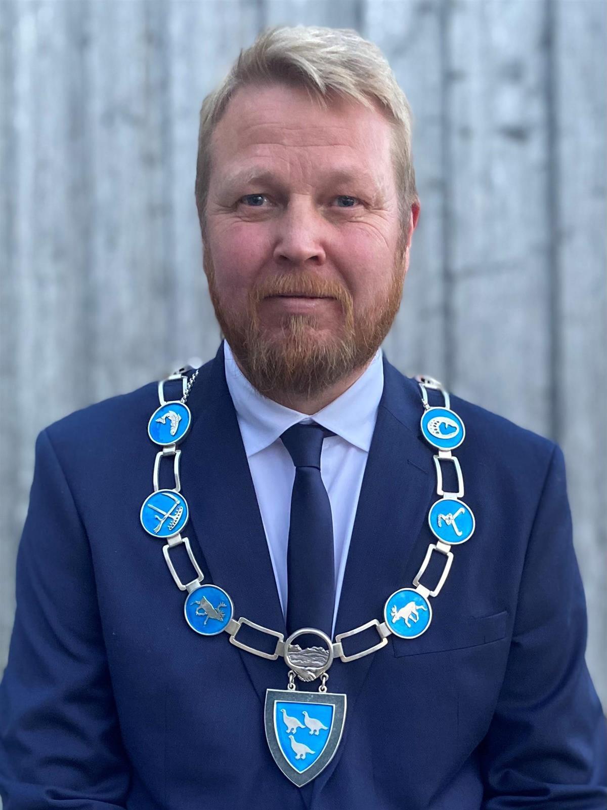 A man in a navy blue suit and tie, wearing a silver and blue ceremonial chain.