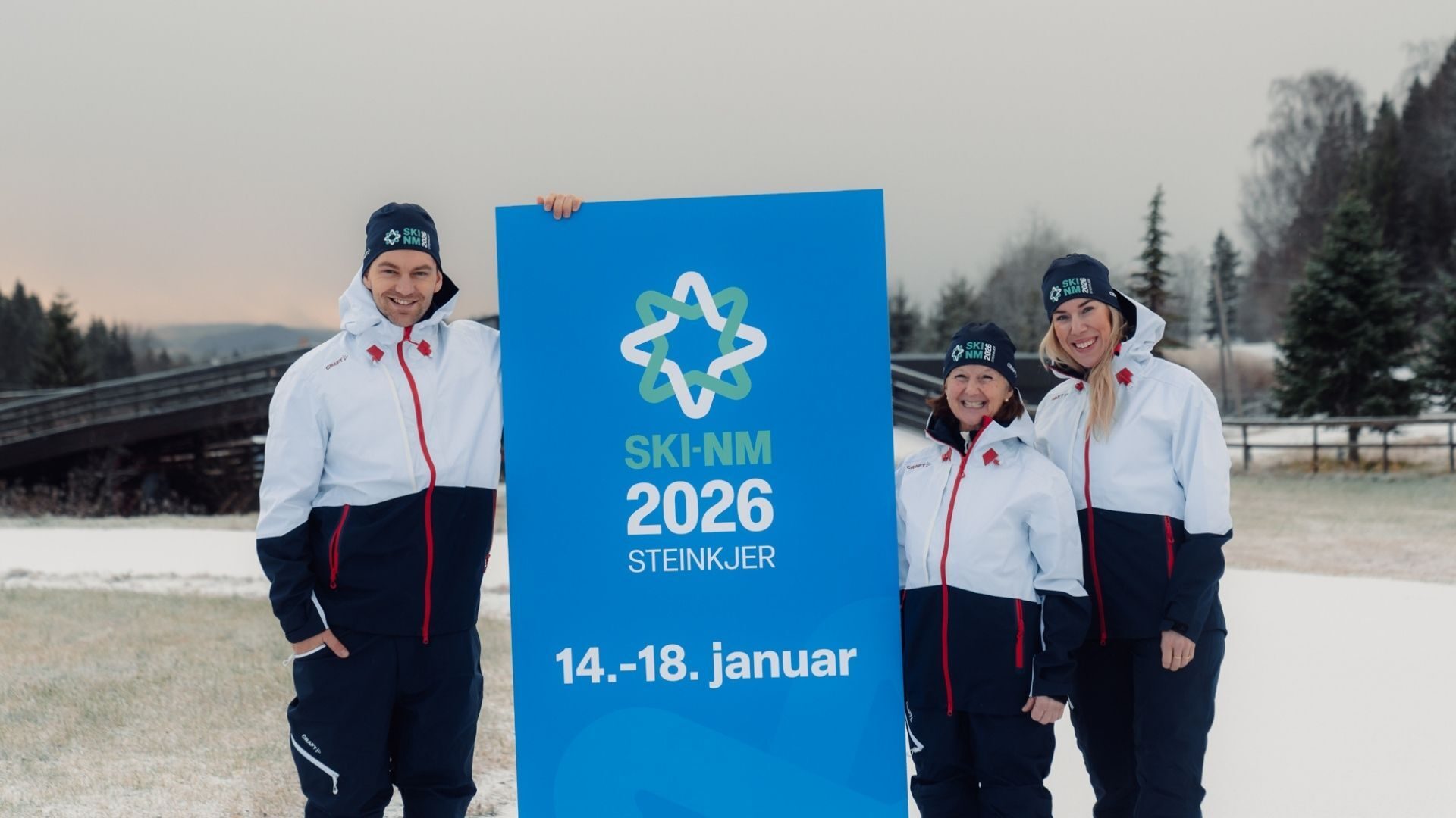 Three smiling people in ski gear hold a blue sign for SKI-NM 2026 Steinkjer in a snowy landscape.
