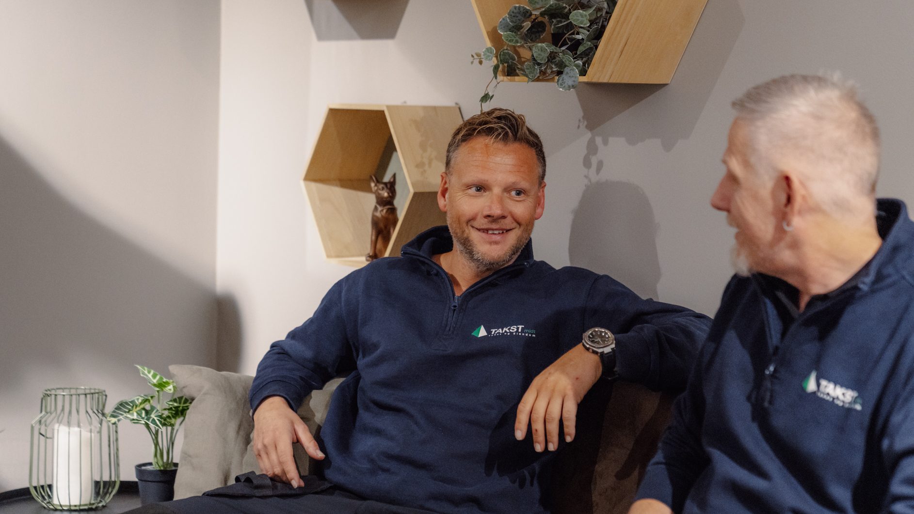 Two men in blue sweatshirts sitting on a couch, one smiling.