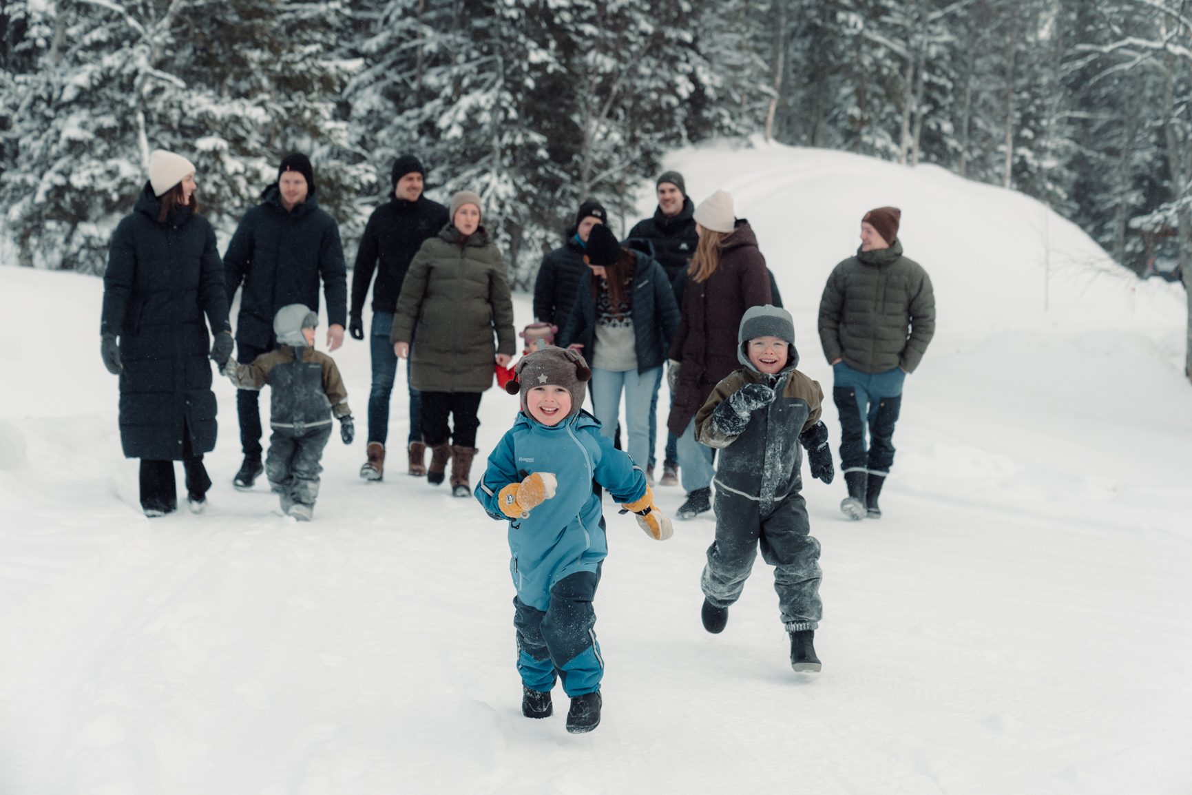 Two young children happily running in the snow, with adults and other children walking behind in a winter forest.
