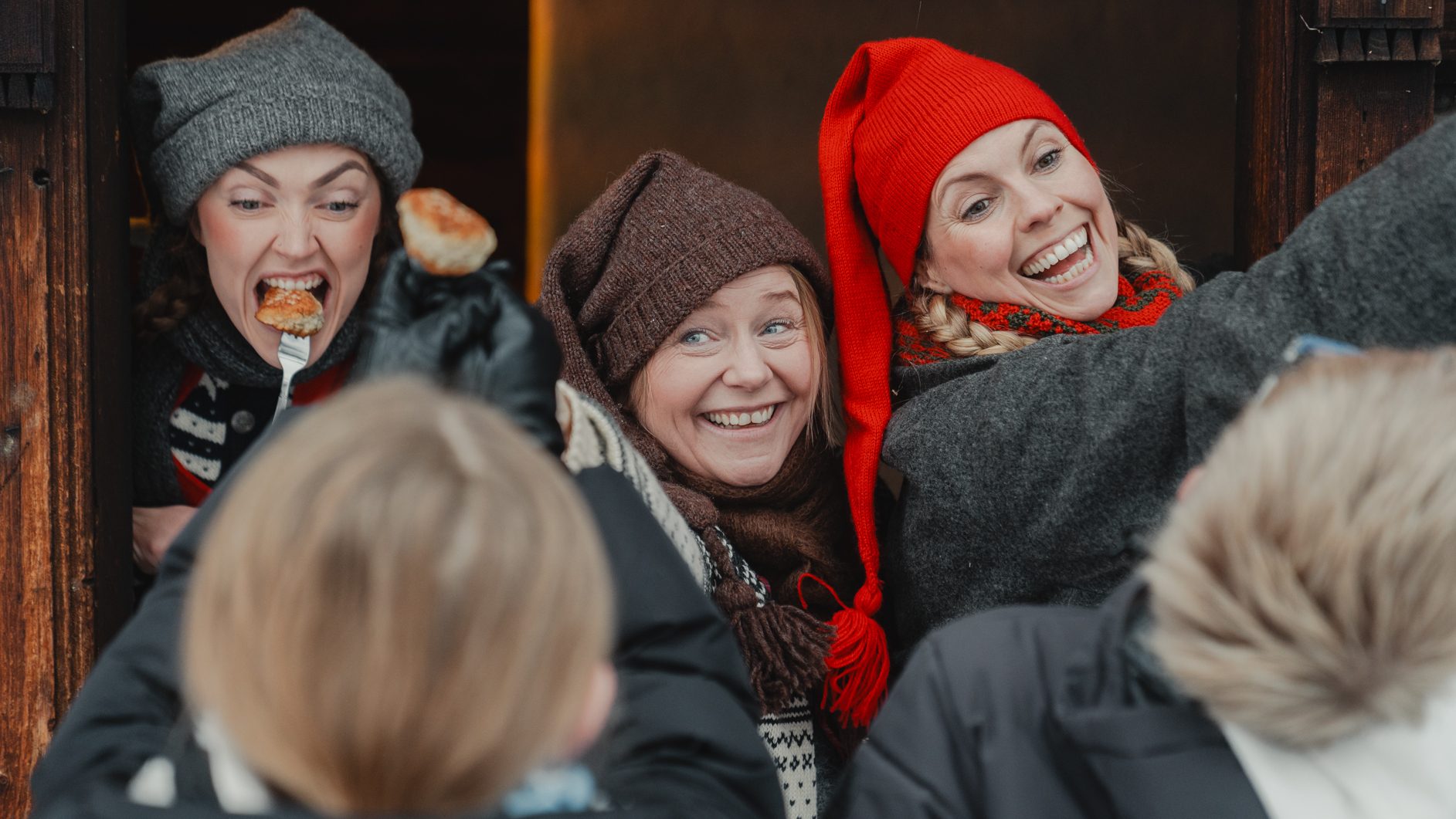 Three women in winter attire, one eating from a fork, two smiling, interacting with blurred children.