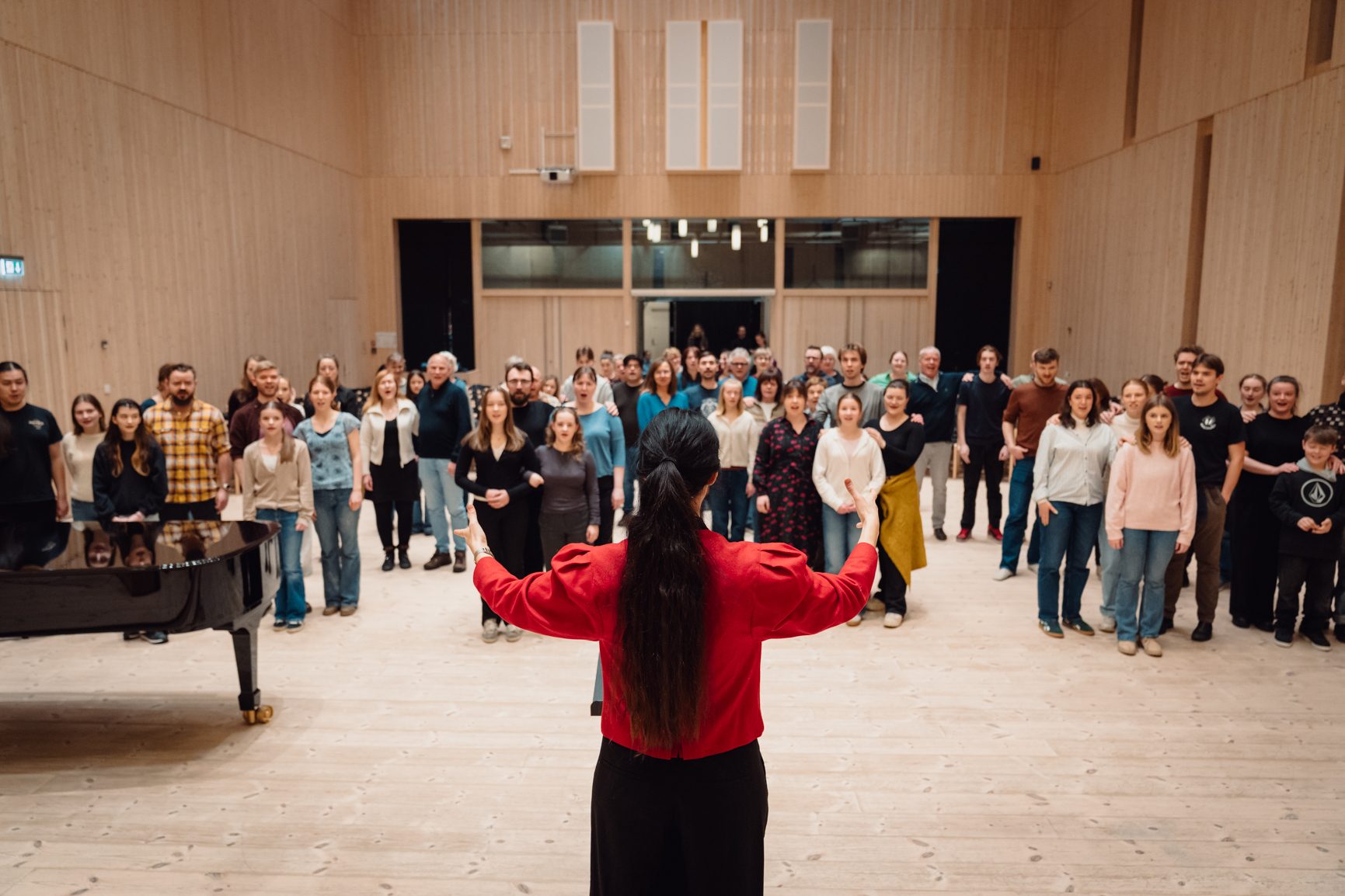 A conductor, back to camera, leads a diverse choir in a spacious wooden hall.