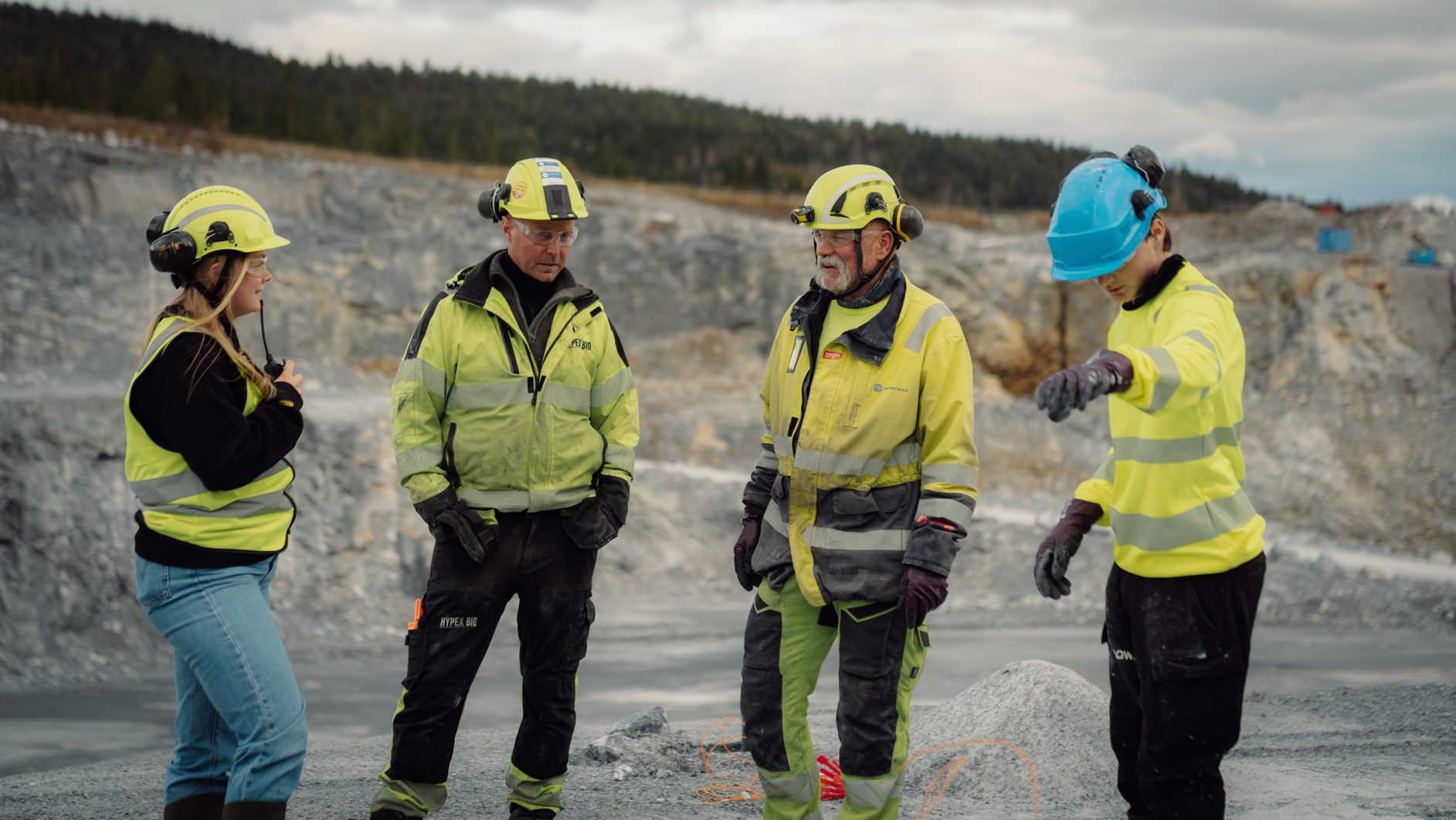 Four workers in hard hats and high-vis gear discussing in a quarry.