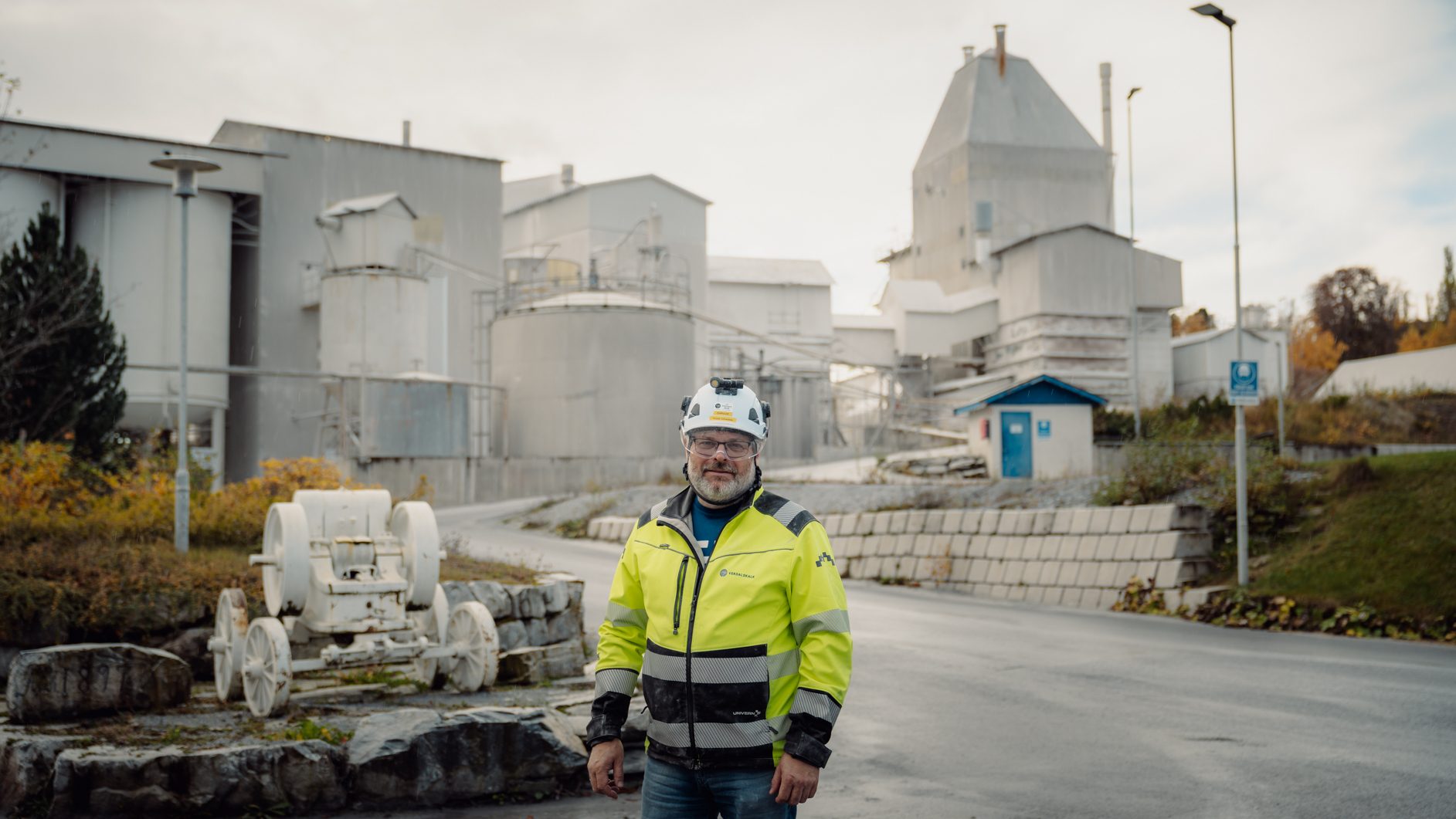 Man in hard hat & hi-vis jacket at an industrial plant with silos and an old cart.