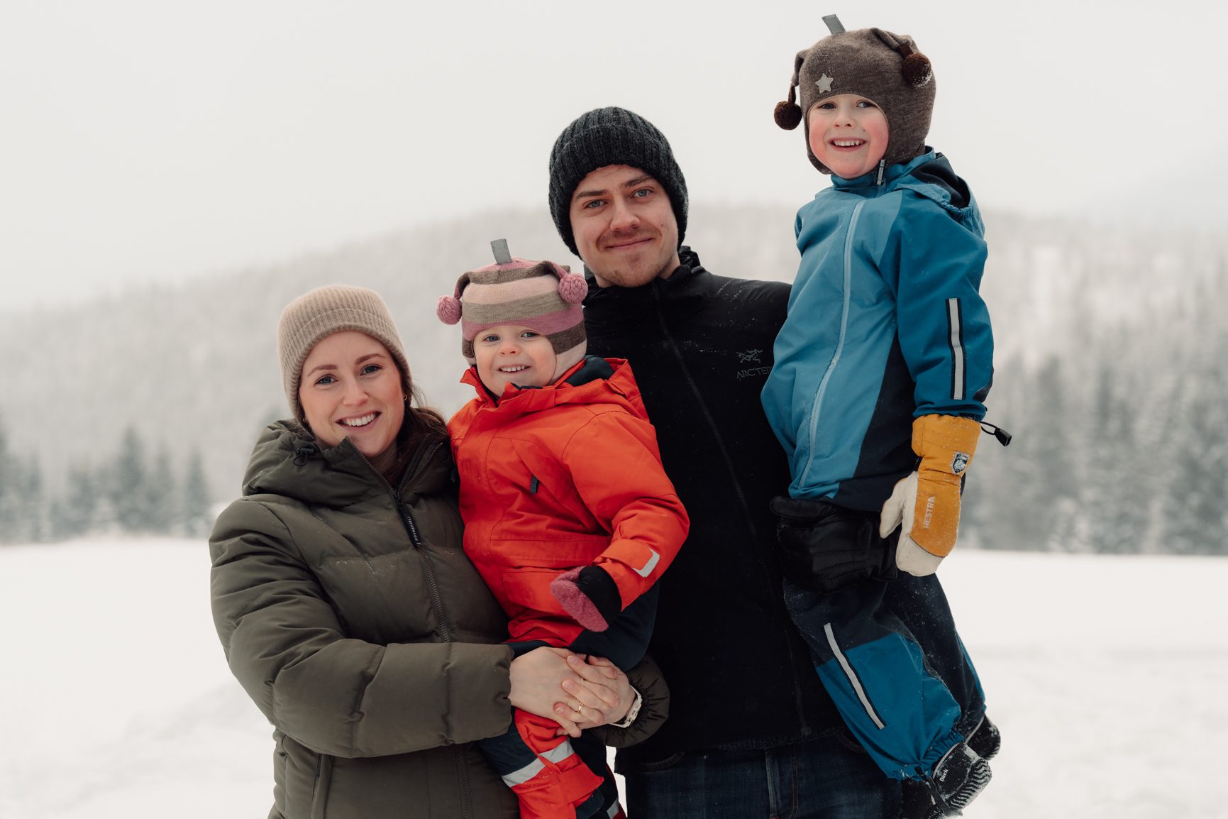 Family of four smiling in winter attire in a snowy landscape.
