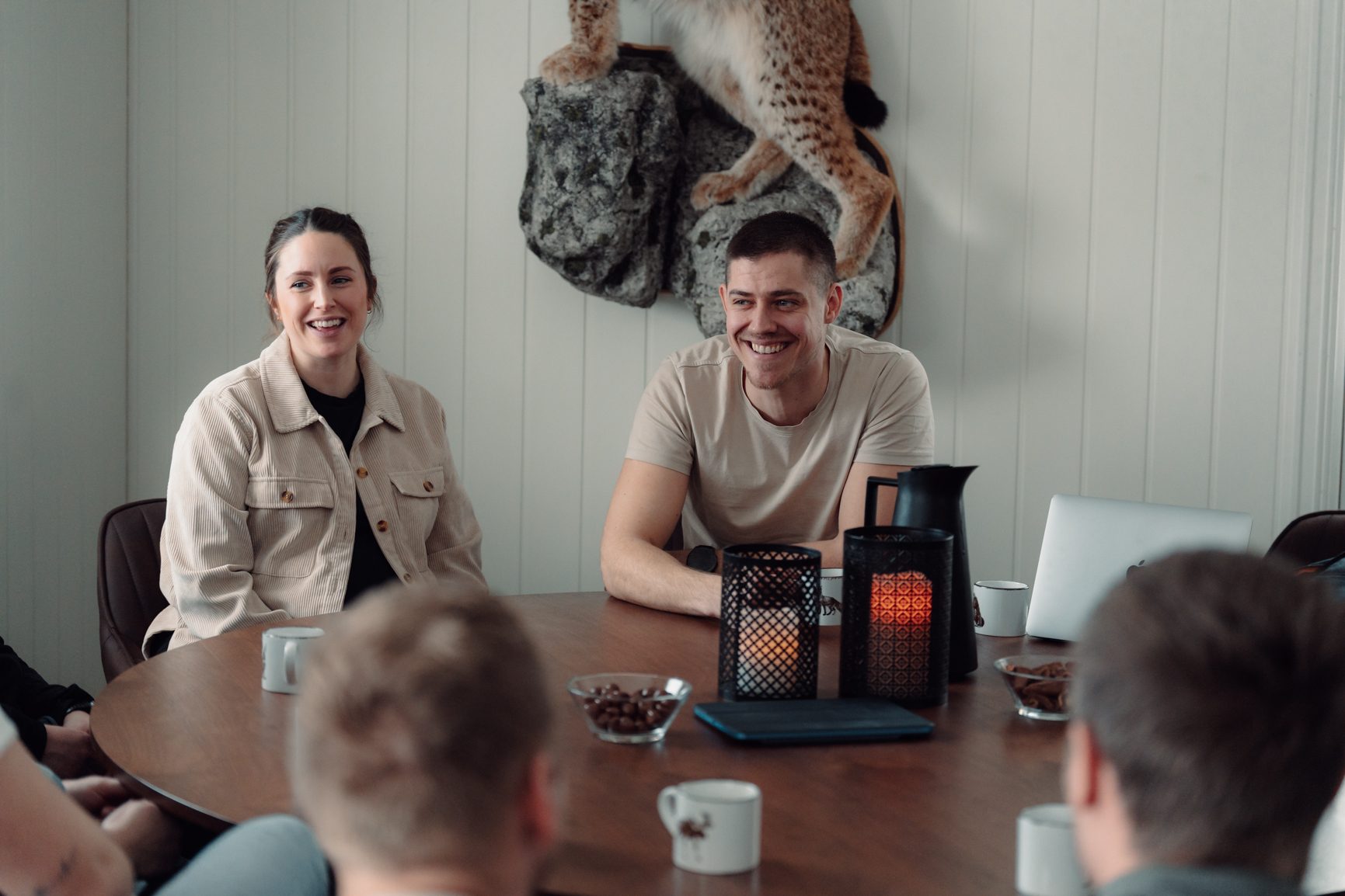 A man and woman smiling at a table with others, coffee, and lanterns in a bright room.