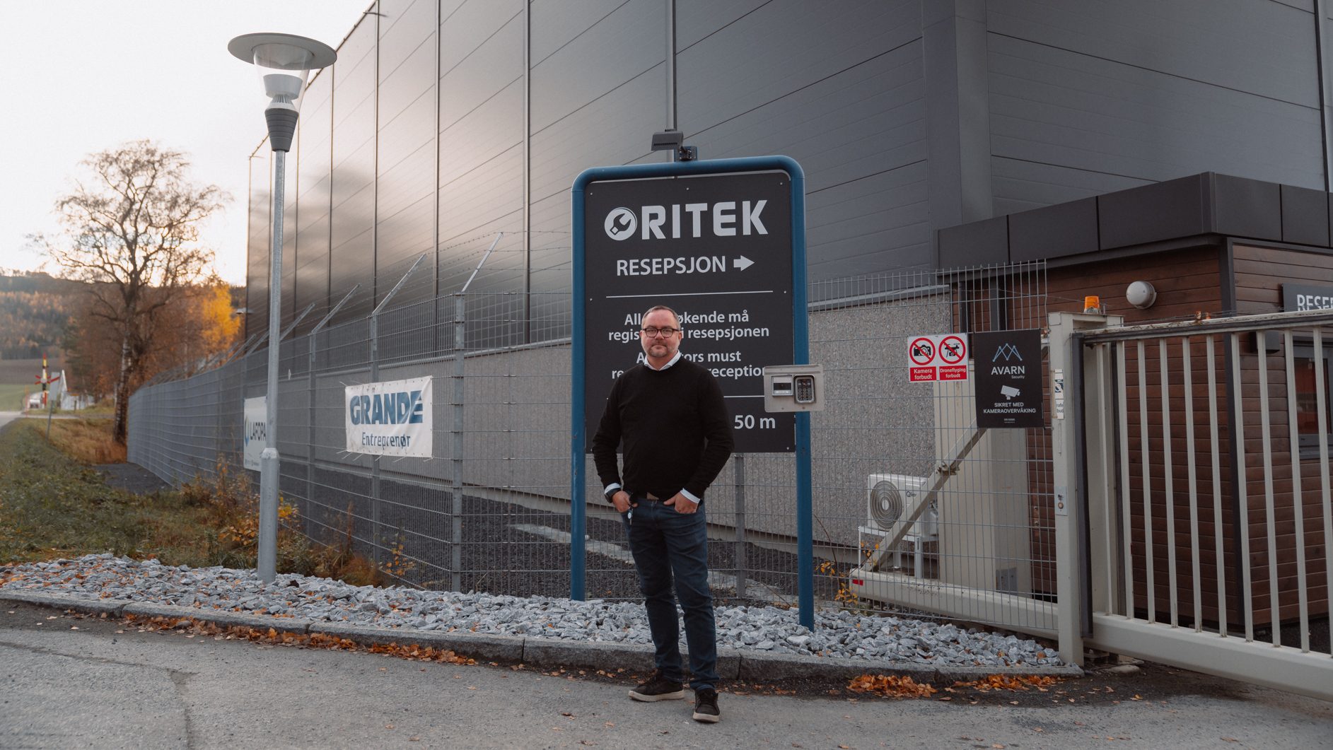 A man stands before a modern industrial building, next to an "ORITEK RECEPTION" sign.