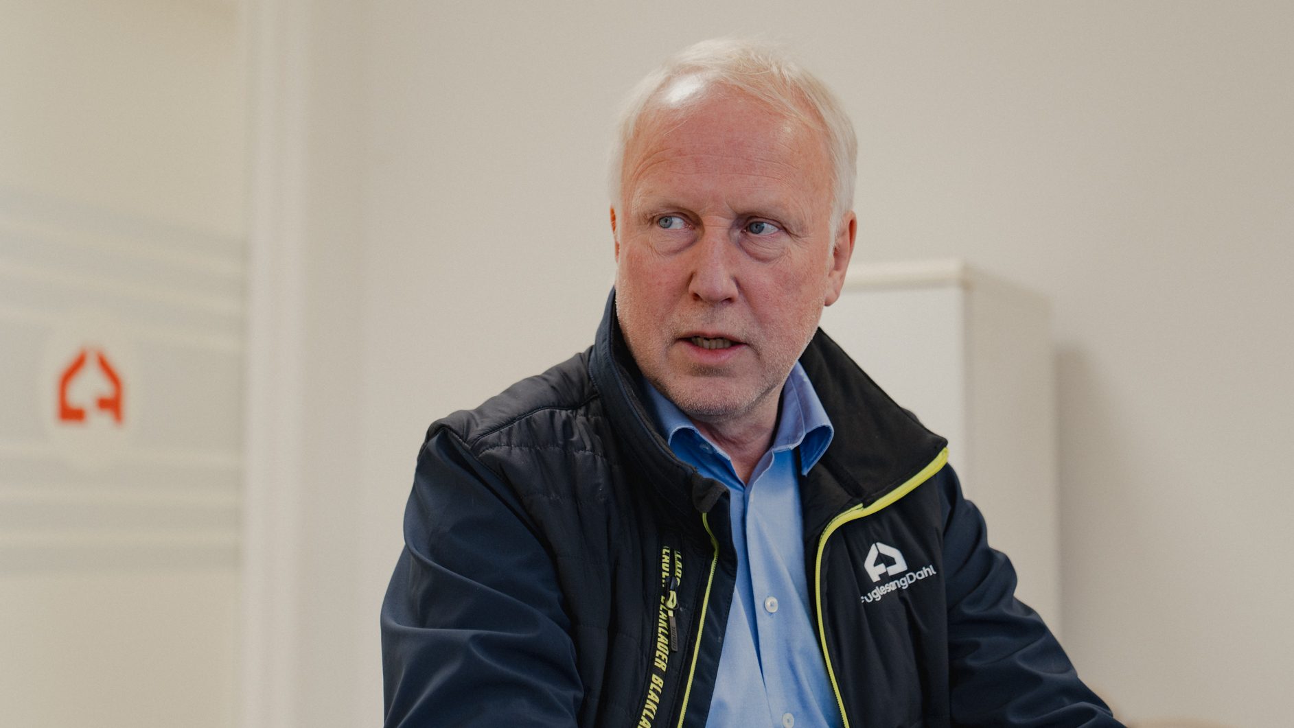 Man with white hair, wearing a vest with a "FuglesangDahl" logo, looking left.