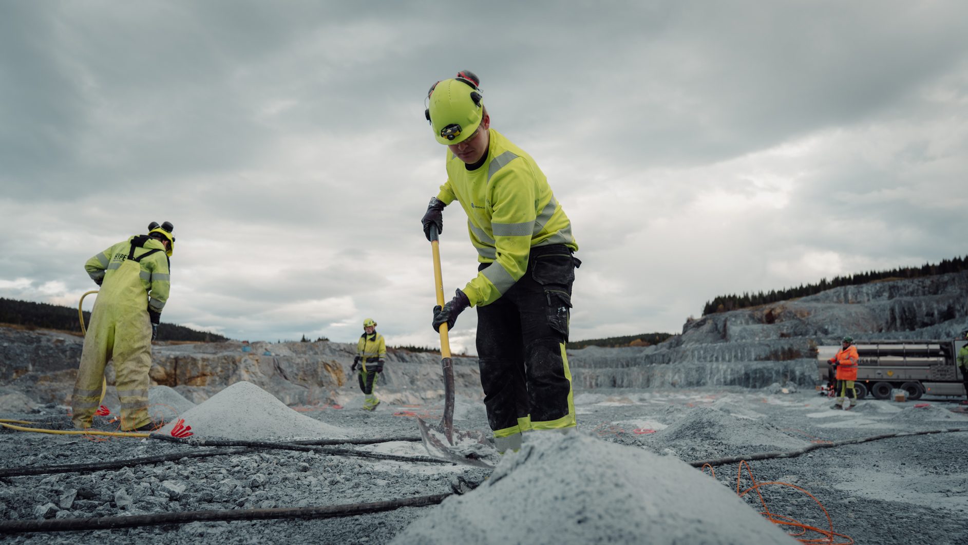 Workers in safety gear shovel gravel at a quarry under a cloudy sky.