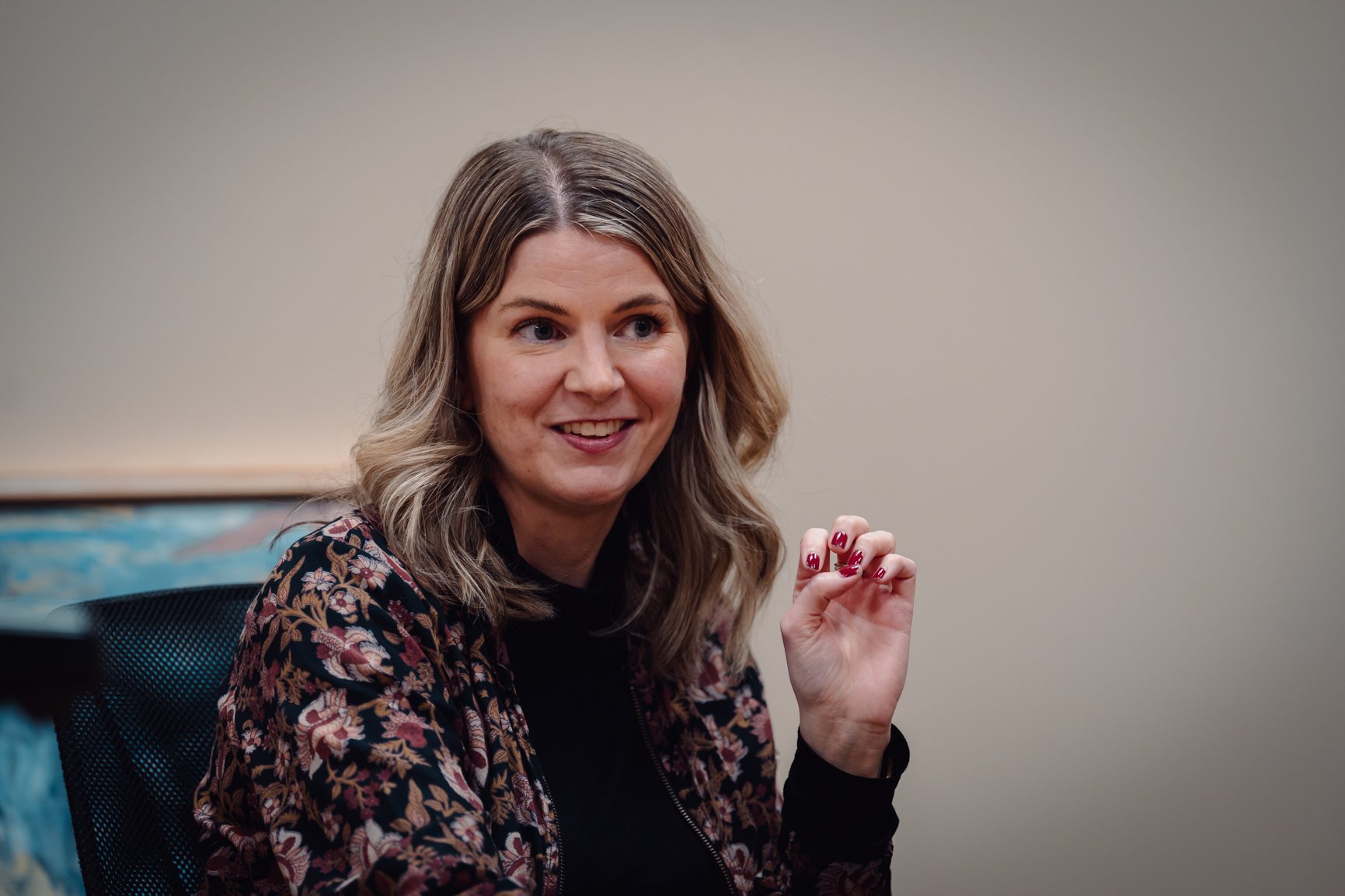 A smiling woman with blonde hair in a floral jacket gestures with her hand, wearing red nail polish.