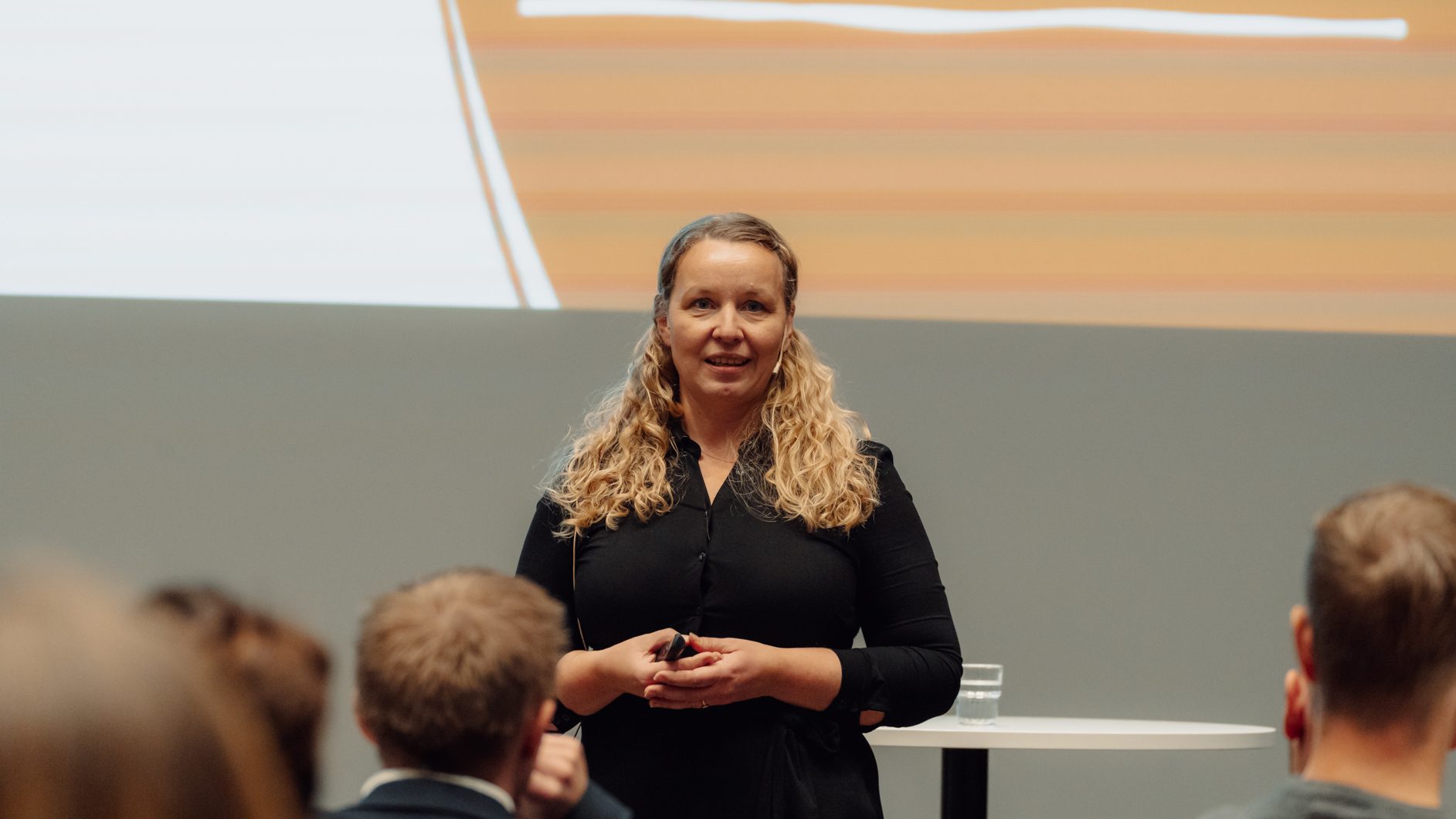 Woman in black shirt presents to audience, holding remote, with screen in background.