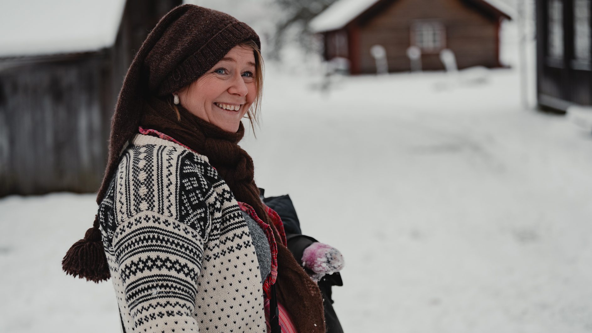 Smiling woman in a cozy winter hat and sweater in a snowy village scene.