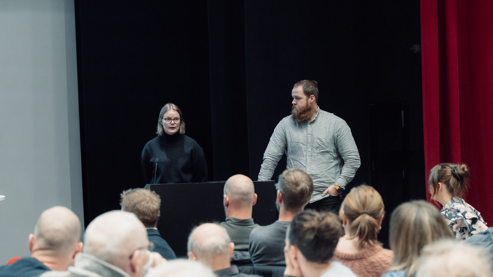 Two speakers (woman and man) address an audience at a presentation.