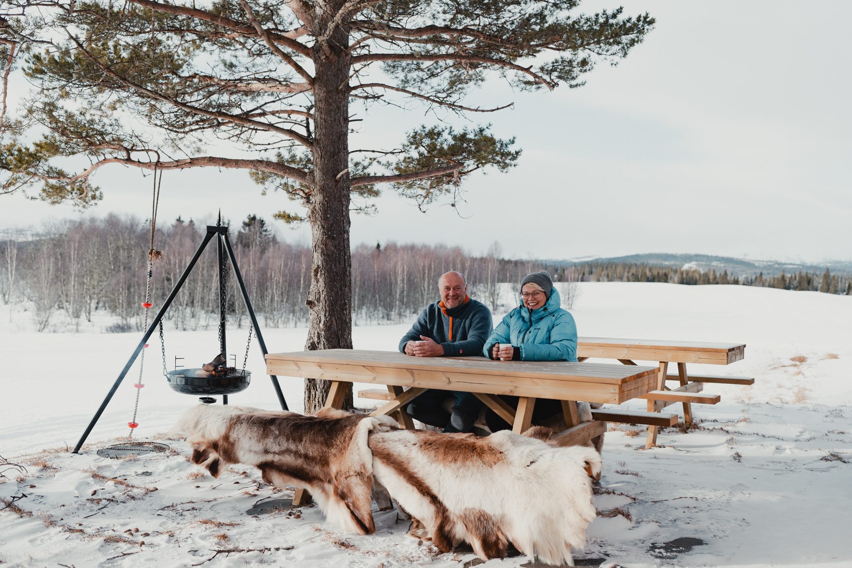 Two people at a snowy picnic table with a tree, fire pit, and animal furs.