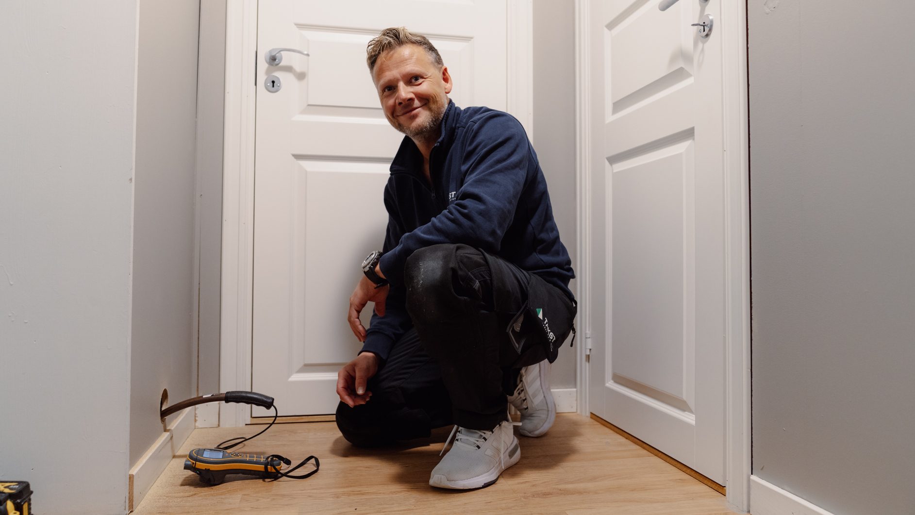 Smiling man kneeling, inspecting a wall with a probe and meter in a hallway.