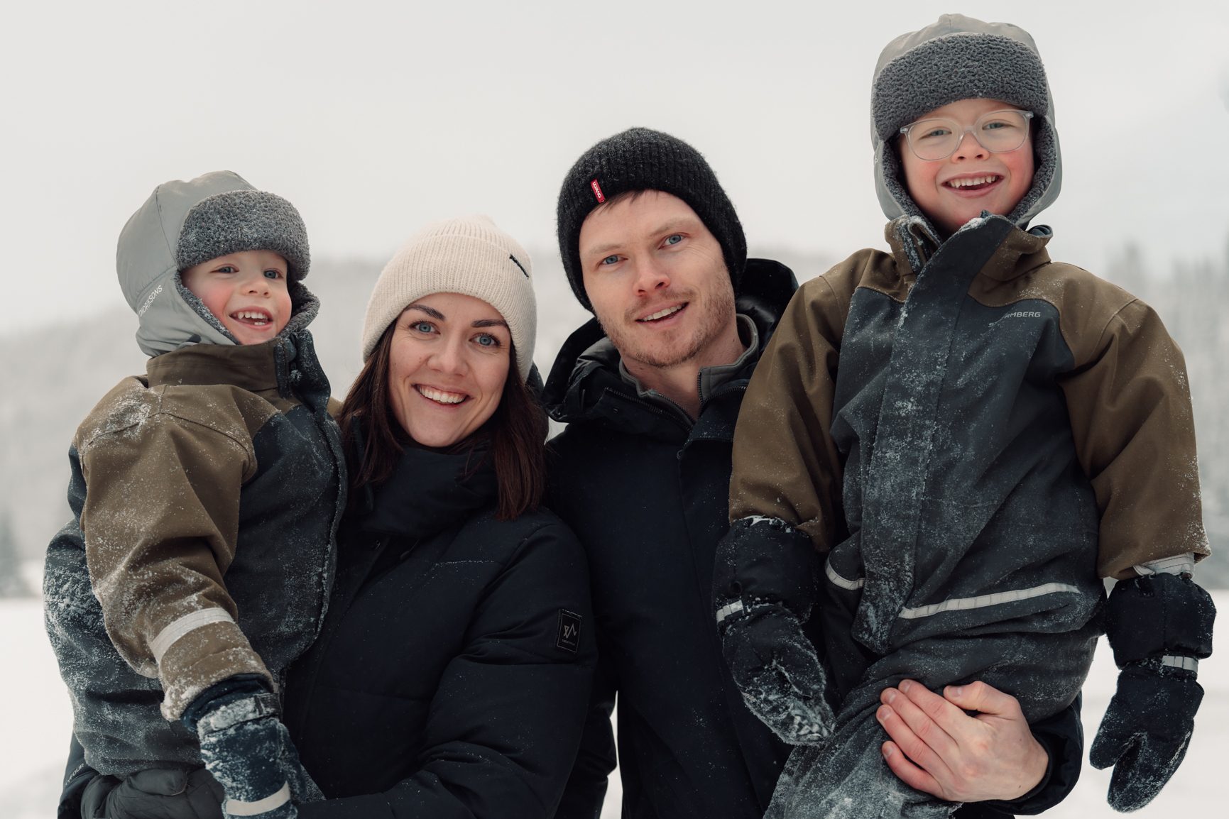 Smiling family of four (two adults, two boys) in winter clothes in snow.