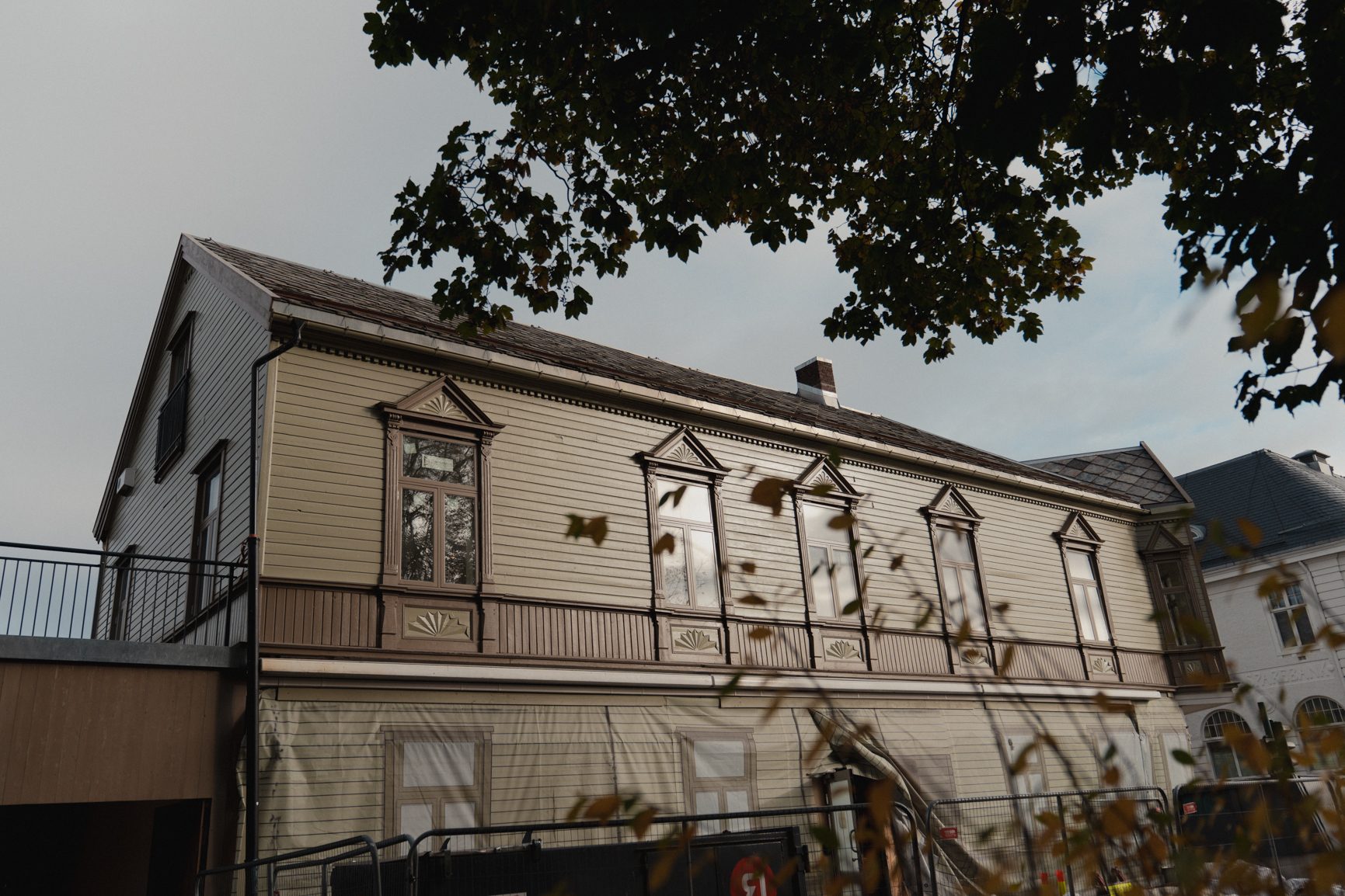 Light brown wooden building with ornate windows and trim, partially seen through dark tree leaves.