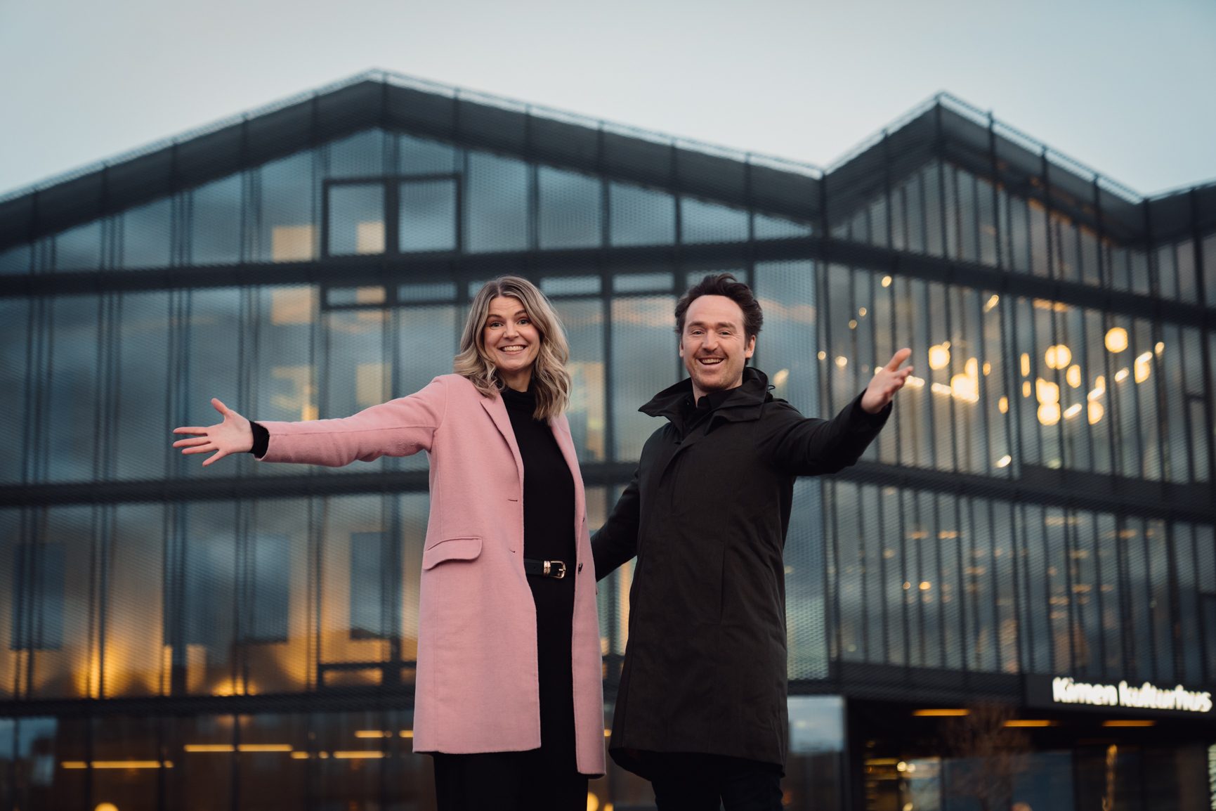 A man and woman in coats smile and gesture in front of a modern glass building.