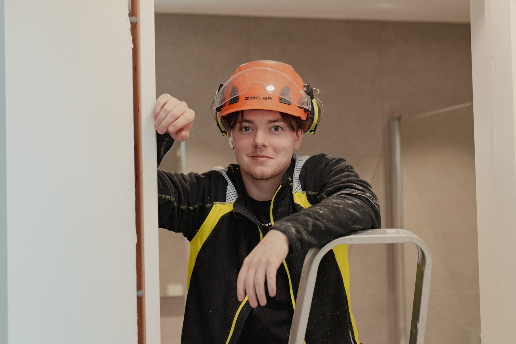 Young man in orange safety helmet and work jacket, leaning on a ladder.
