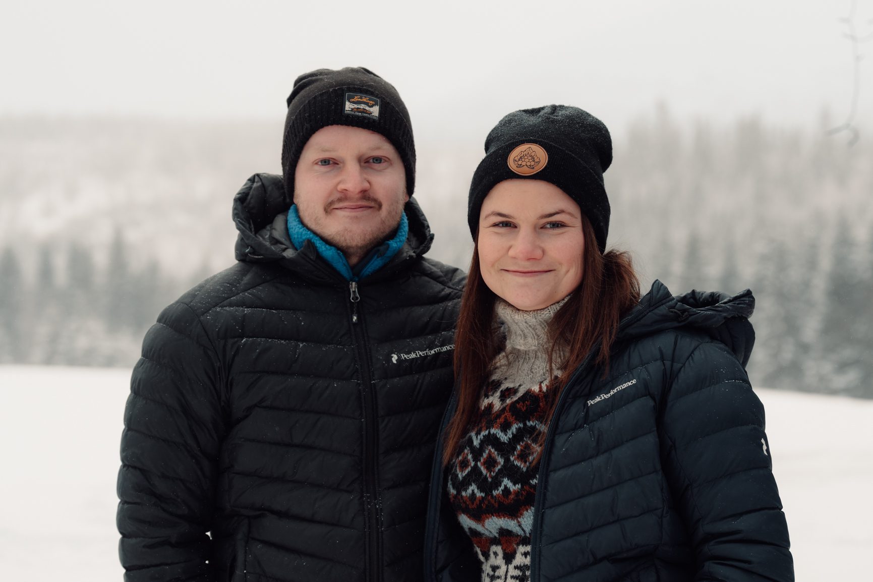 Two people in winter jackets and beanies stand side-by-side in a snowy outdoor landscape.
