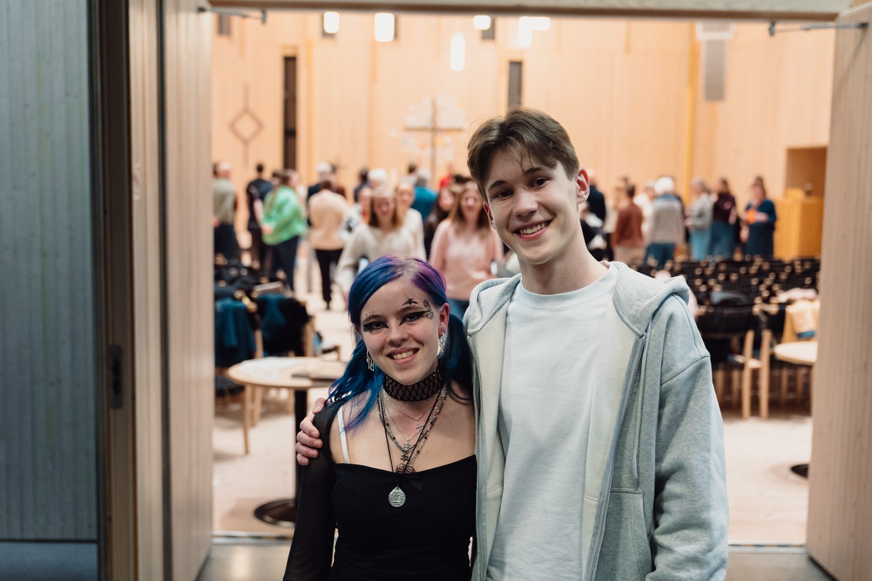Two smiling young people, a girl with blue hair and a boy in a hoodie, standing in a large hall.