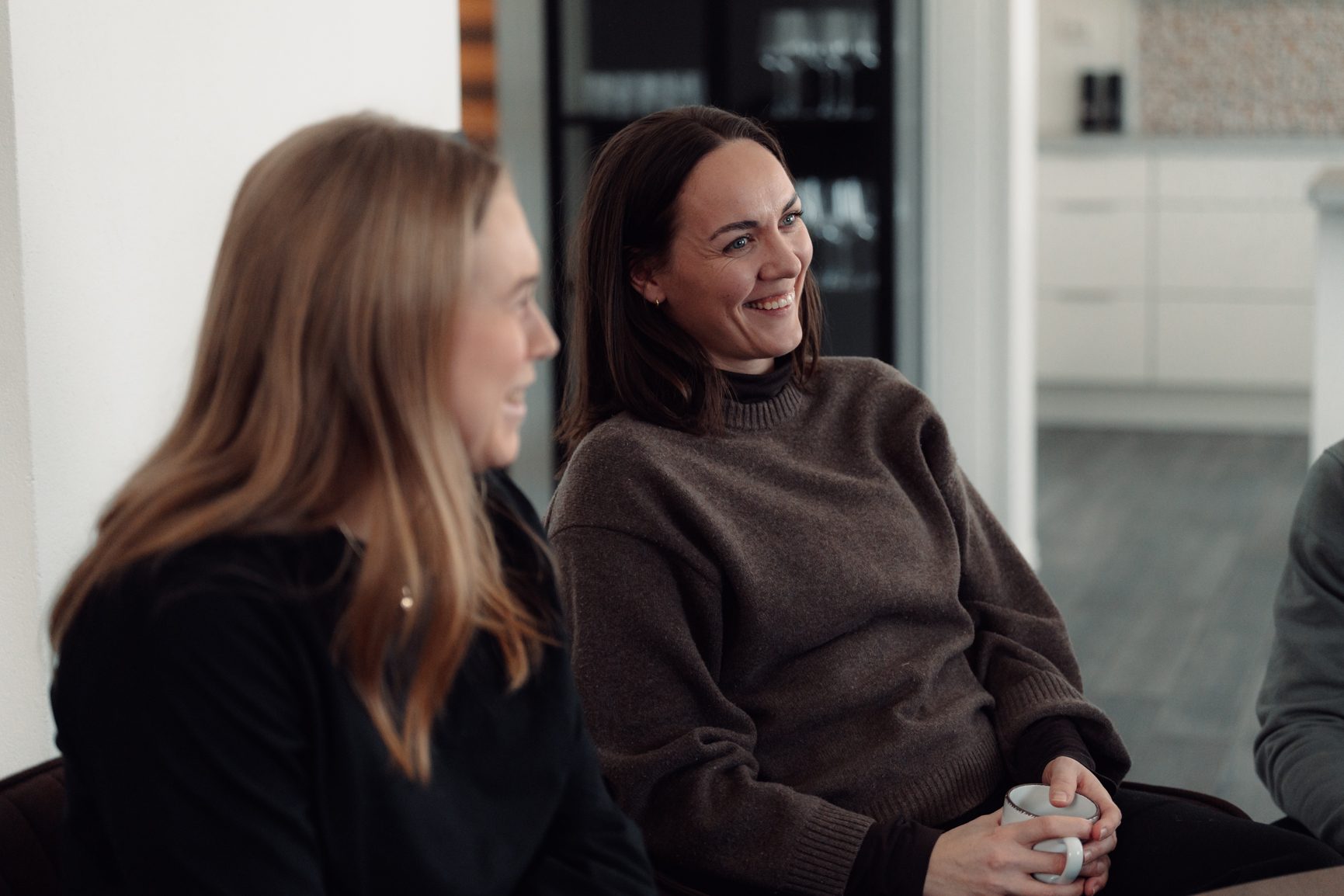 Two women in conversation, one smiling and holding a mug, in a bright indoor setting.