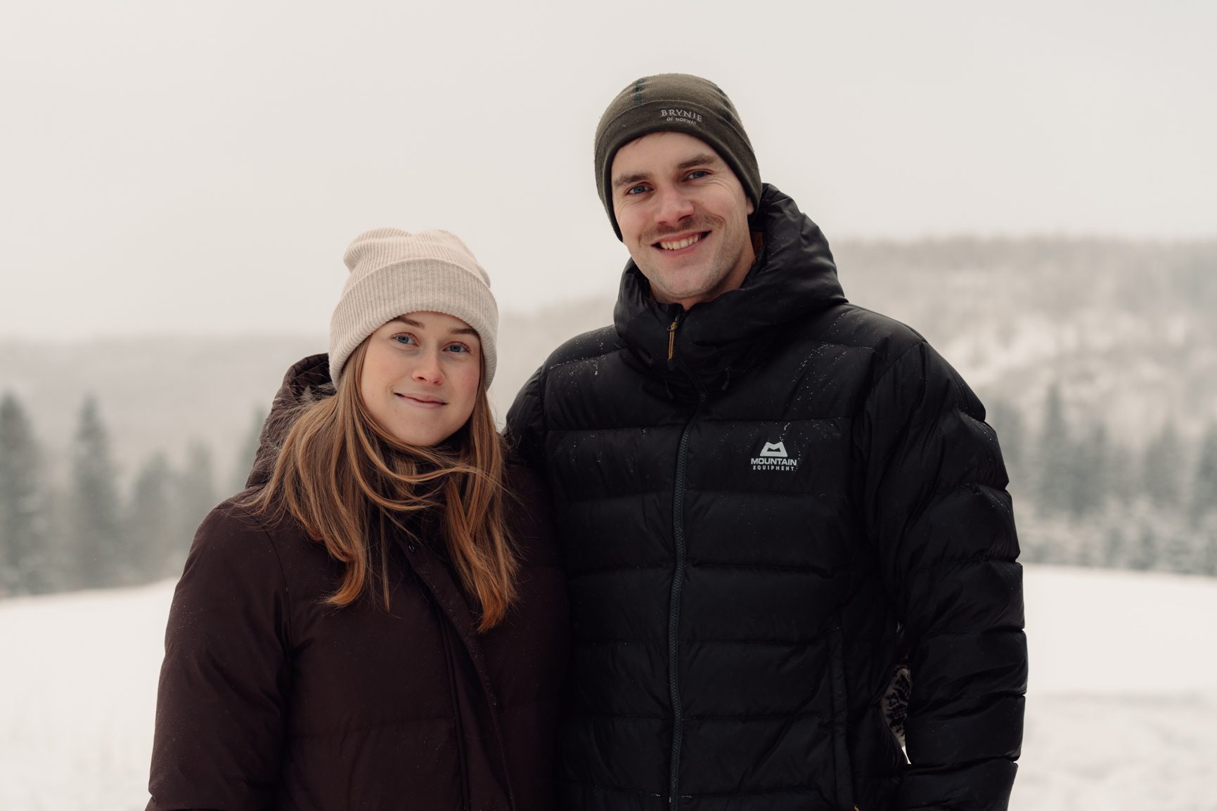 A smiling man and woman in winter jackets and beanies stand in a snowy, outdoor landscape.