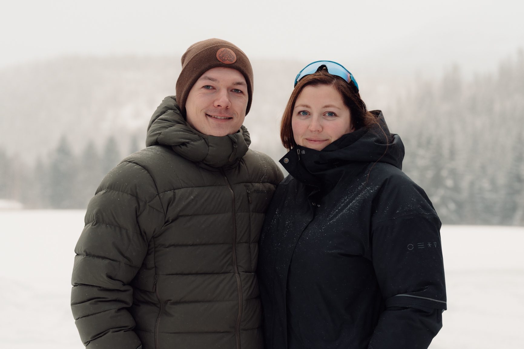 Two smiling people in winter coats and hats standing in a snowy landscape with trees in the background.