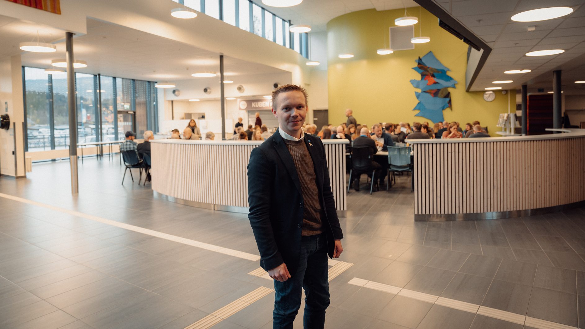 A man in a jacket stands in a bright, modern hall with people dining and large windows in the background.