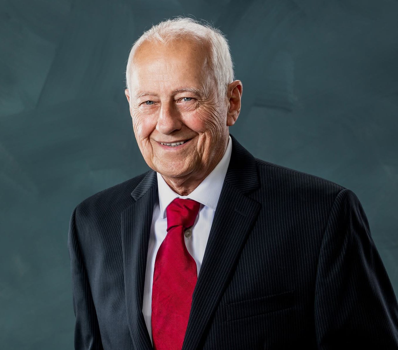 A smiling elderly man with white hair in a suit and red tie.