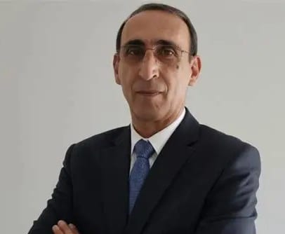 Man in a dark suit, light blue tie, and glasses, standing with arms crossed, looking at the camera.