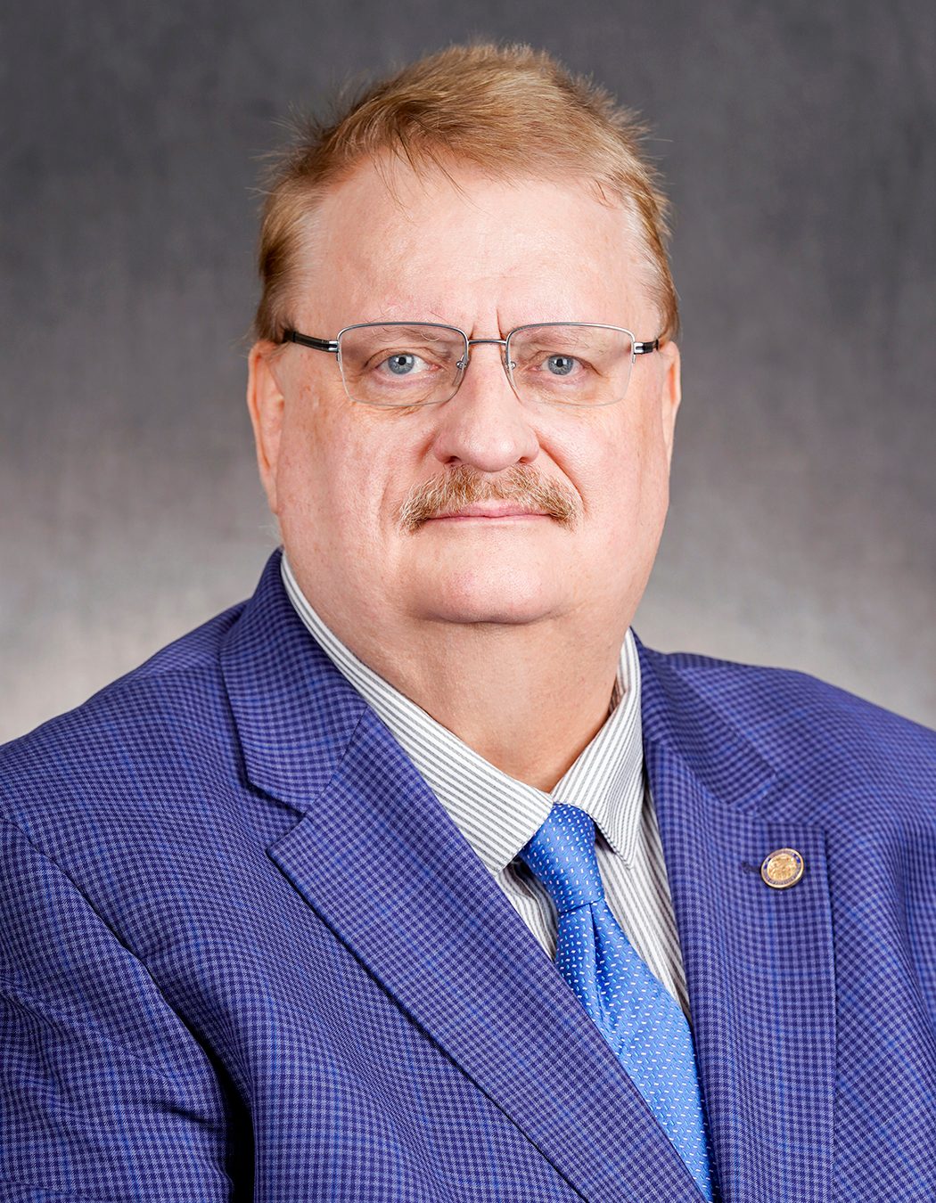 Headshot of a man with glasses and a mustache, wearing a blue checkered suit, striped shirt, and blue tie.
